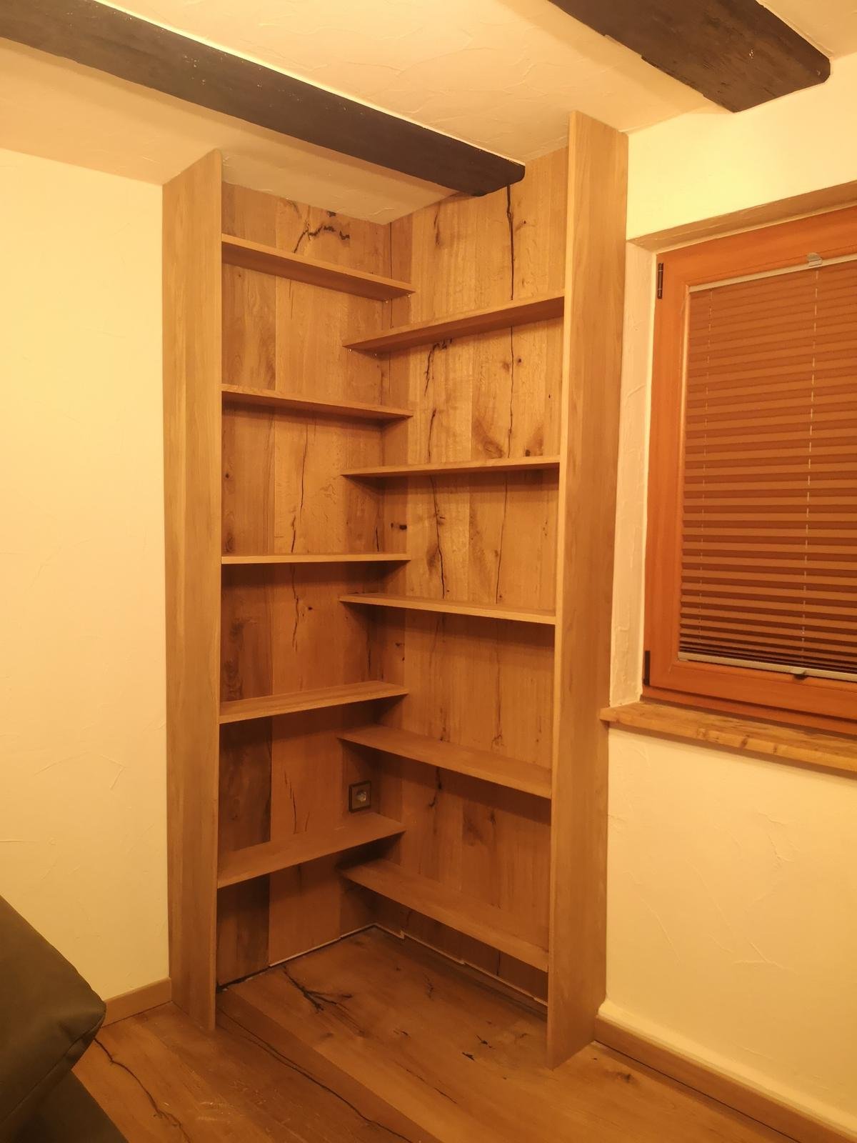 Ein Holzregal mit mehreren Regalbflächen, eingebaut in eine Ecke eines Zimmers, neben einem Fenster mit Holzrahmen.