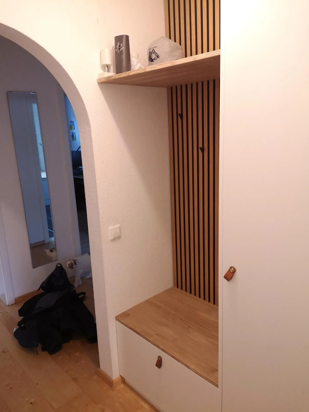 Ein Eingangsbereich mit einem weißen Schrank, einer offenen Holzablage, einer vertikalen Holzlamellenwand und einer oberen Holzablage. Im Hintergrund sieht man eine Tür mit einem Spiegel, einige Mäntel und eine Katze am Boden.