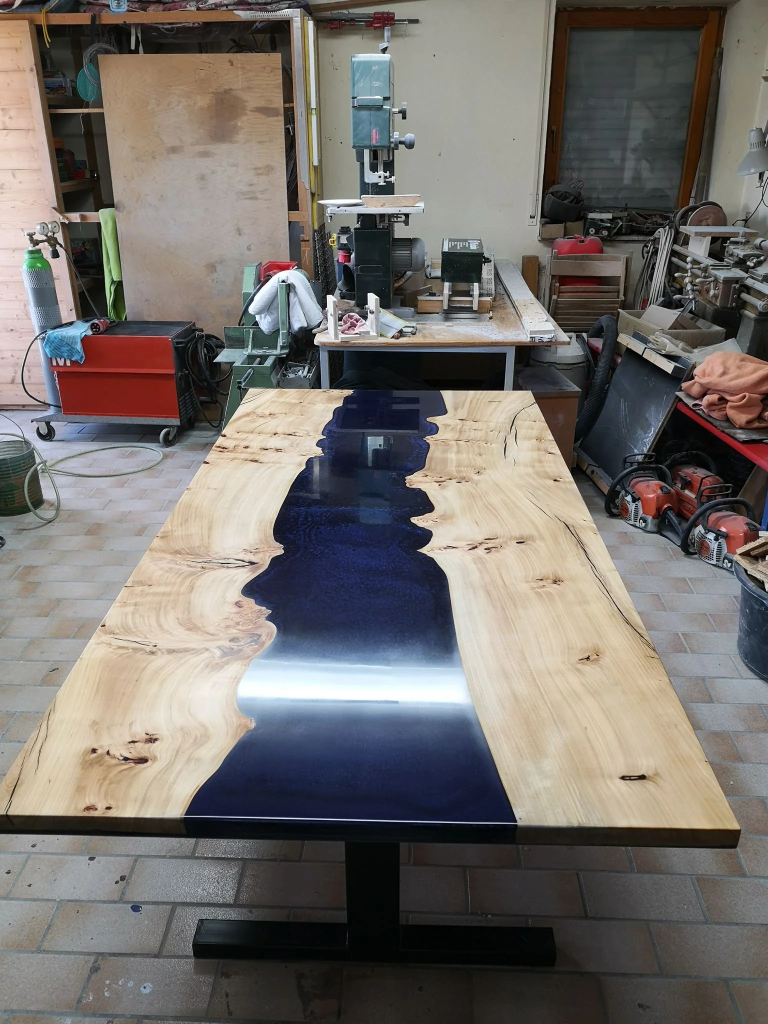 Ein handgefertigter Holztisch mit schwarzem Fluss in der Mitte, in einer Werkstatt mit Werkzeugen und Maschinen im Hintergrund.