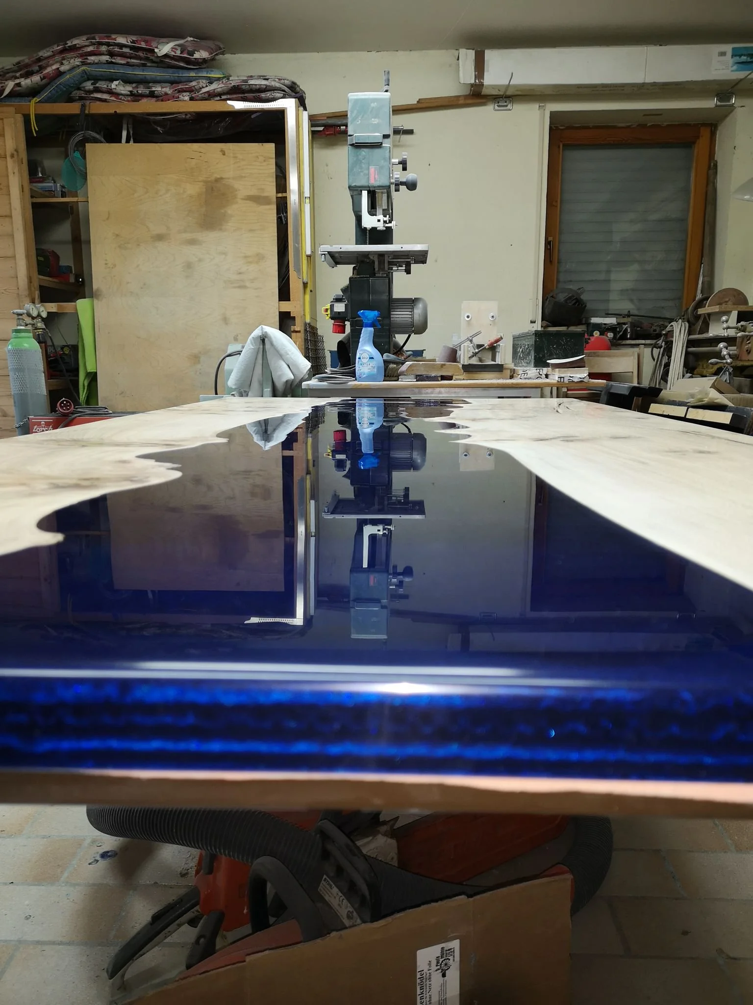 Ein Arbeitstisch mit einer lackierten Holzplatte, die im Raum eines Werkzeug- oder Holzverarbeitungsraums liegt, im Hintergrund Maschinen und Werkzeuge.