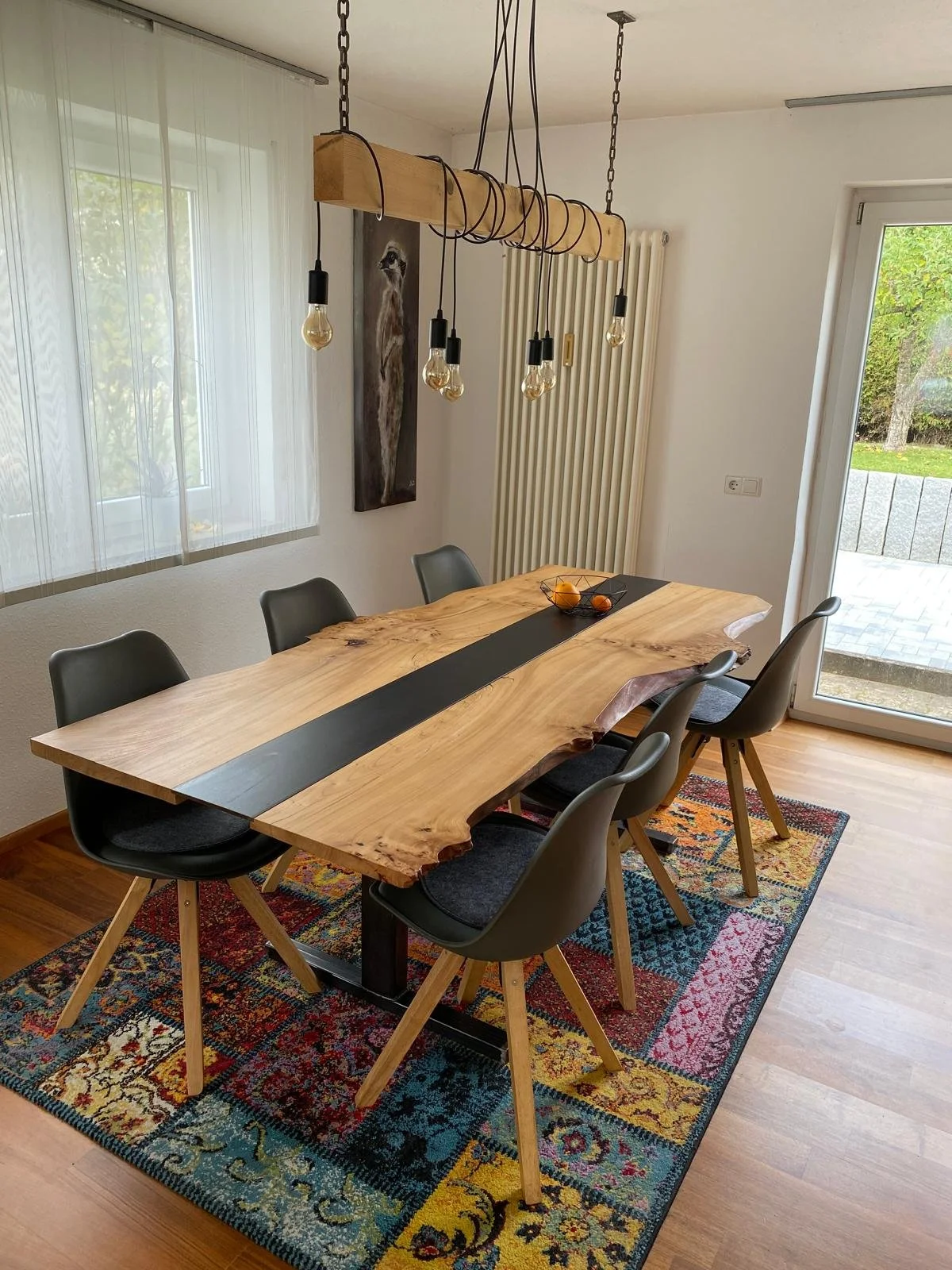 Moderner Esszimmertisch mit sechs schwarzen Stühlen, dekorative Pendelleuchte über dem Tisch, farbenfroßer Teppichboden, Wandbild eines Tieres, große Fenster und Tür nach draußen.