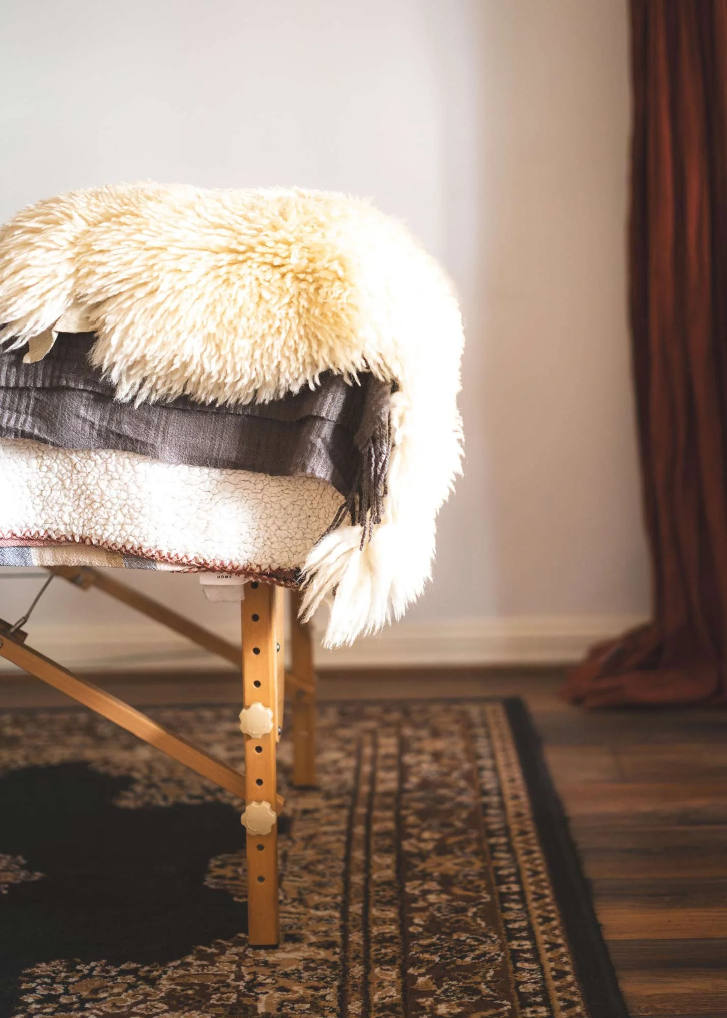 A wooden stool with a fluffy cream-colored blanket and a dark brown cloth draped over it, placed on a patterned rug next to a wooden floor and a wall with a curtain.