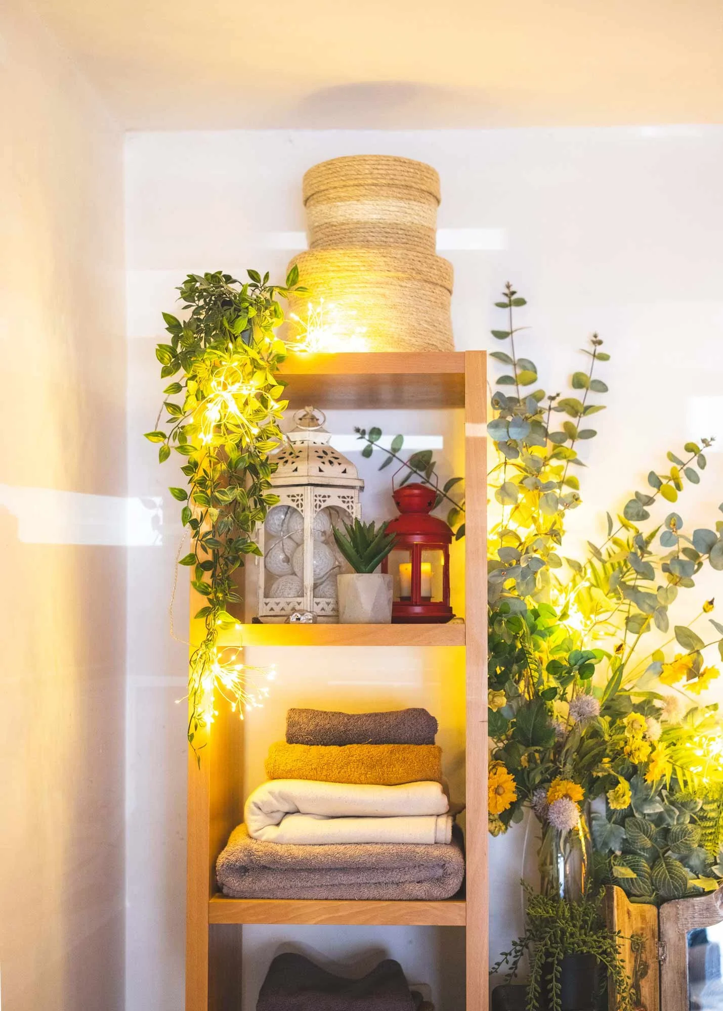 A wooden shelf decorated with plants, books, and decorative items, with string lights, surrounded by green foliage and a large vase of flowers.