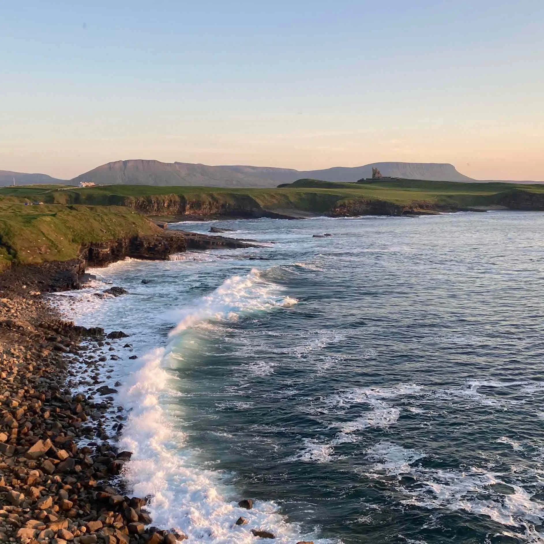 A coastal landscape with rocky shoreline, green cliffs, and hills in the distance, with a calm ocean and gentle waves during sunset.
