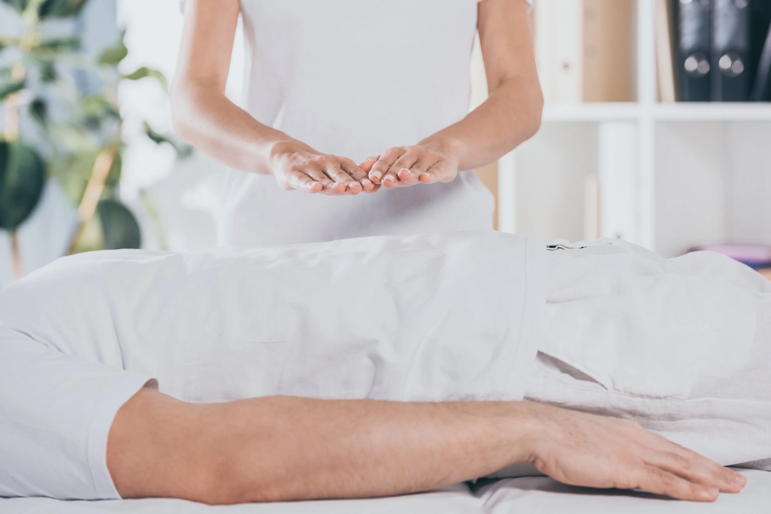 A person in white clothing receiving a massage from another person in a bright, airy room with plants and shelves in the background.