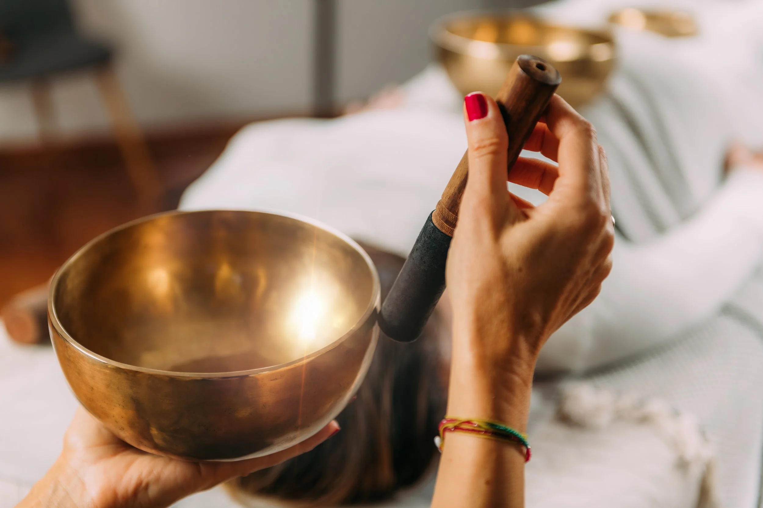 Person holding a singing bowl with a wooden mallet, in a peaceful setting with a bed and decorative bowls in the background.