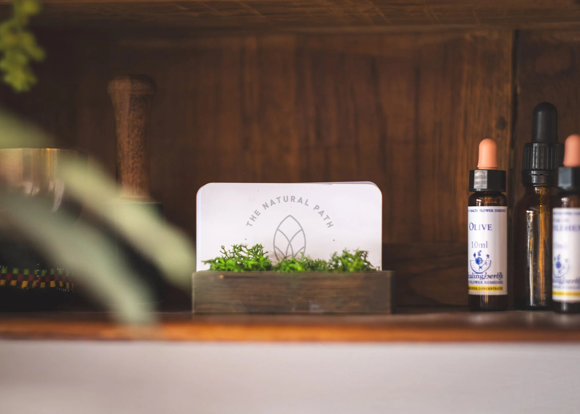 A wooden shelf holding small bottles with dropper caps and a small box with green moss and a label that reads 'The Natural Path' and an illustration of a leaf.