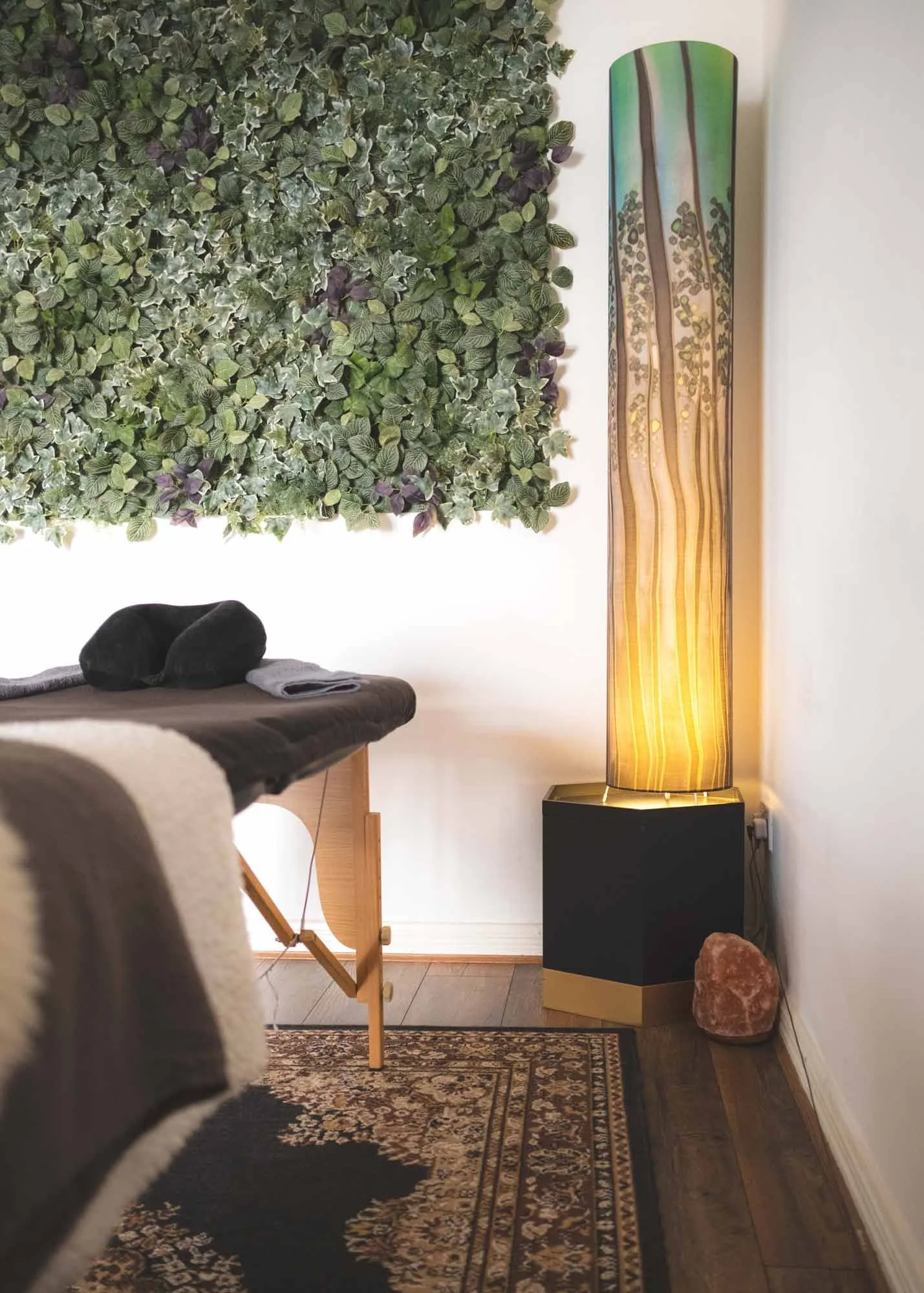 A massage room with a plush massage table, a black towel, and a folded gray towel, against a green leafy wall decor. A tall, colorful, flower-patterned floor lamp stands beside a salt rock on the floor, next to a black and gold storage box. Partial view of a patterned rug and a chair in the foreground.