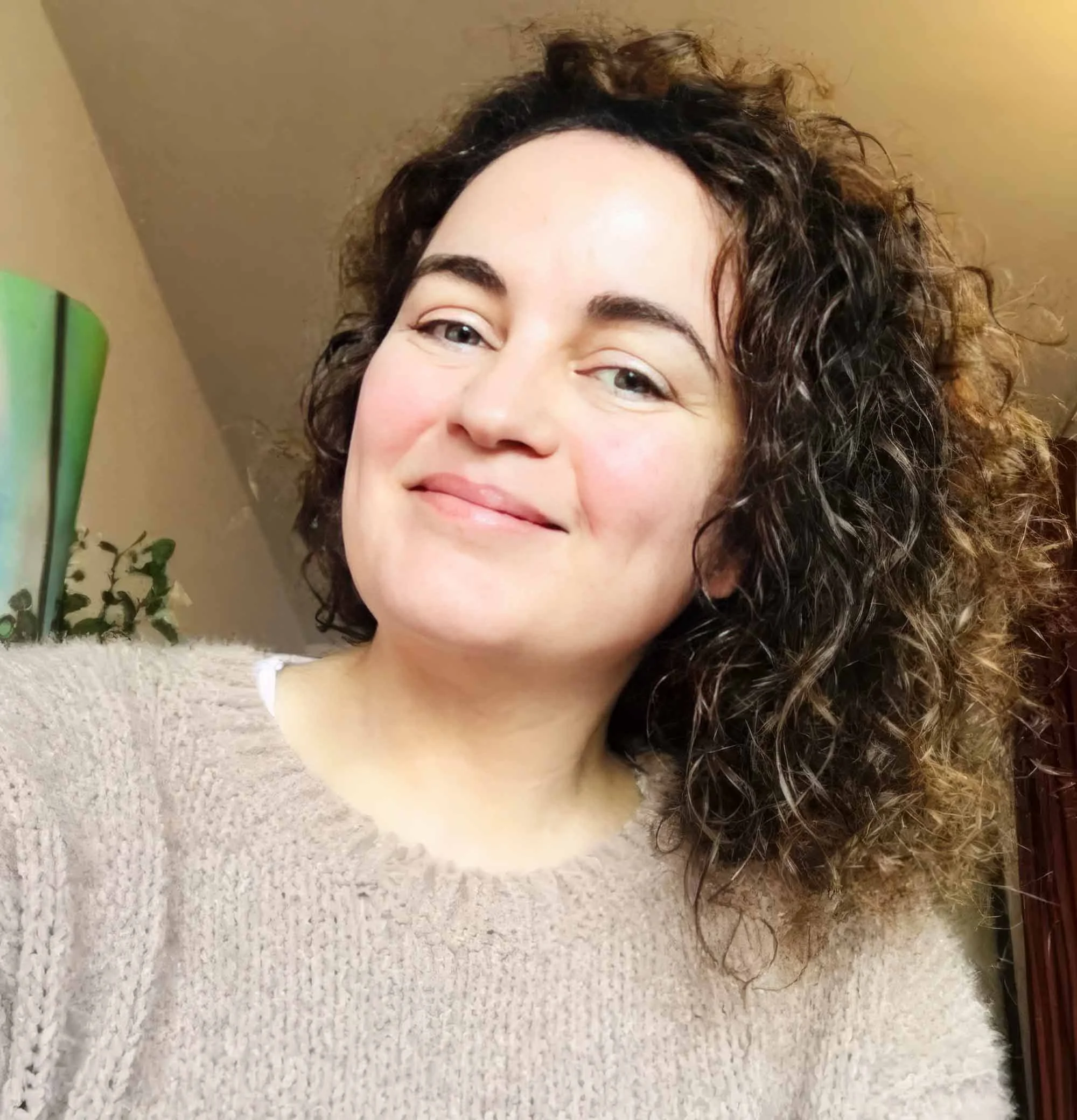 A woman with curly dark hair, light skin, and a gentle smile is taking a selfie indoors, wearing a beige knit sweater.
