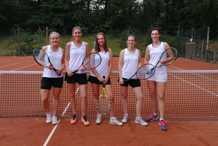Fünf Frauen im Tennisoutfit stehen auf einem Tennisplatz mit Tennisschlägern in der Hand.