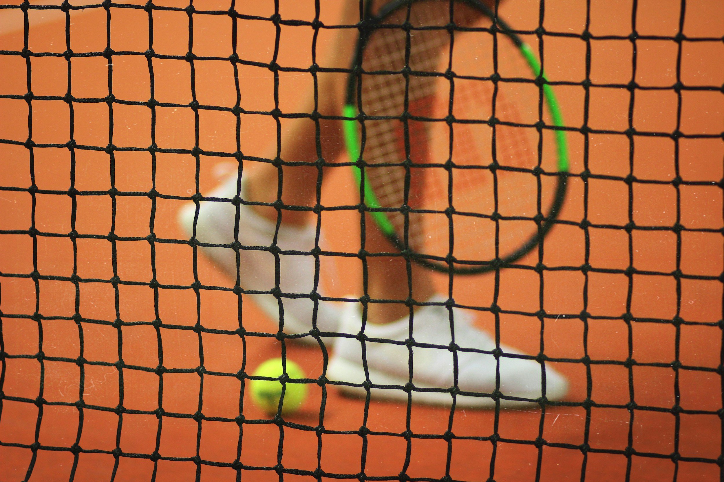 Blick durch ein Tennisnetz auf einen Tennisball, einen Tennisschläger und eine Person auf einem Tennisplatz.