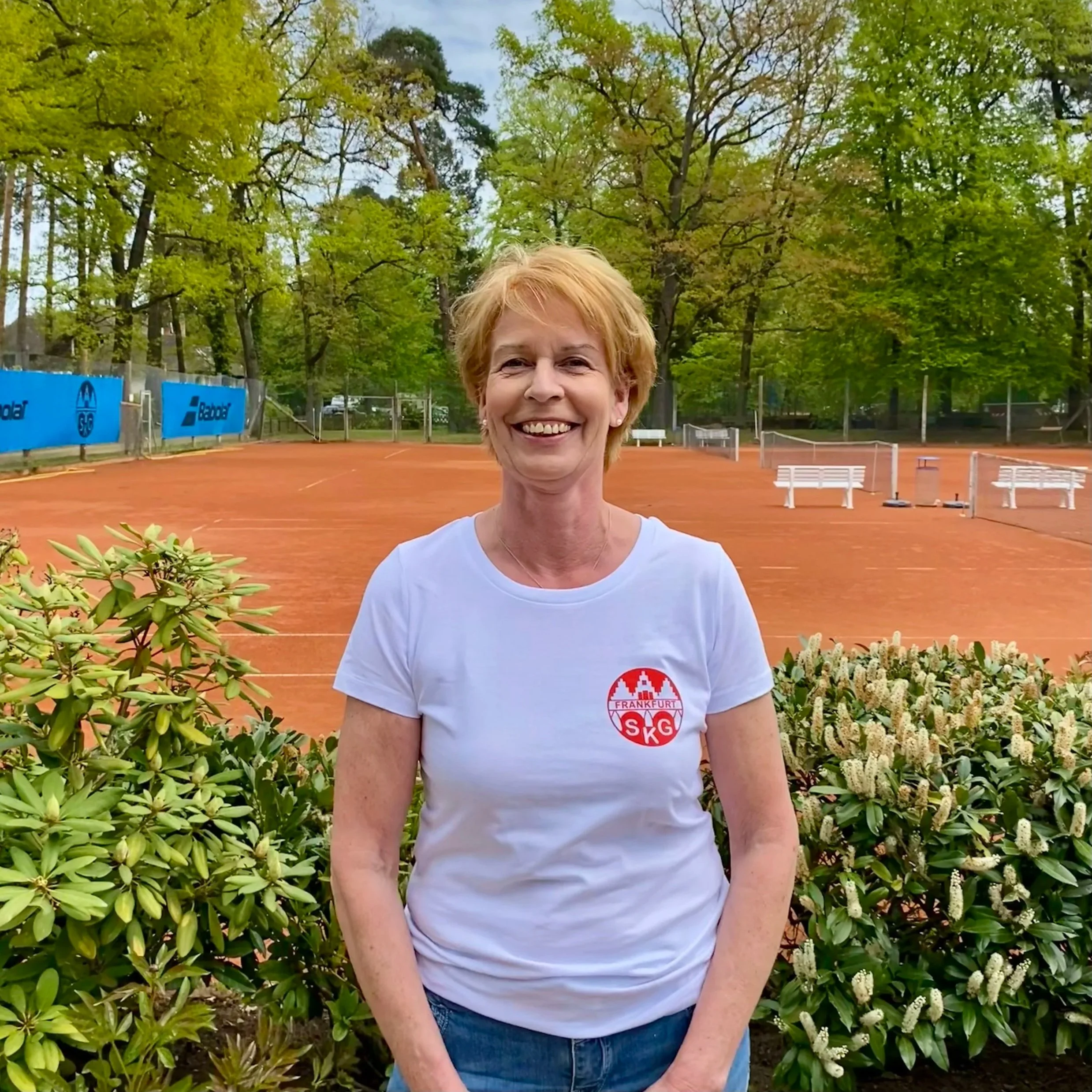 Lächelnde ältere Frau steht vor einem Tennisplatz mit Blumen im Vordergrund, trägt ein weißes T-Shirt mit einem Logo, grüne Bäume im Hintergrund.