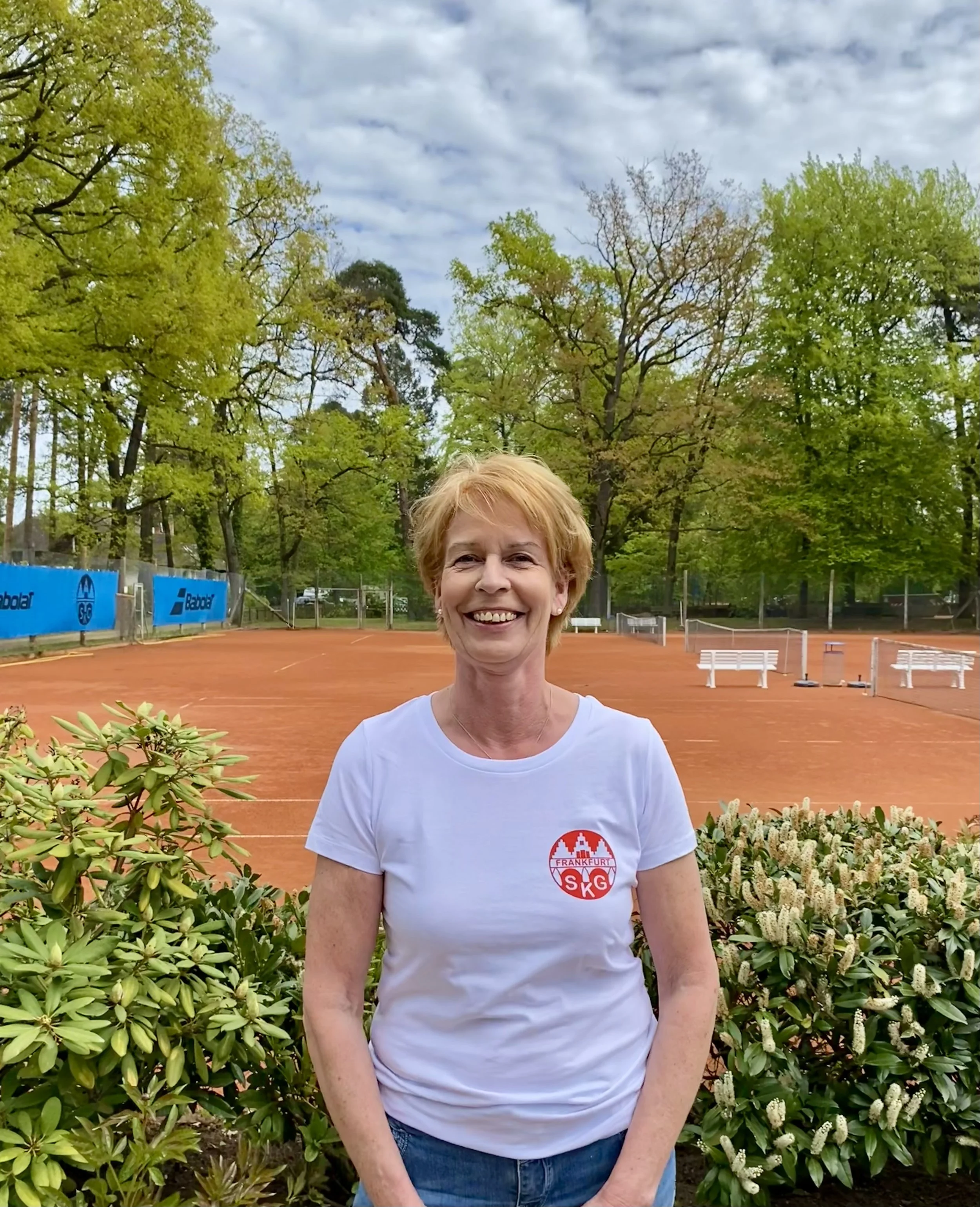Eine lachende ältere Frau trägt ein weißes T-Shirt mit einem roten Logo, steht vor einem Tennisplatz im Freien, umgeben von grünen Büschen und Bäumen, im Hintergrund sind Spielbälle und Tennisnetz sichtbar.