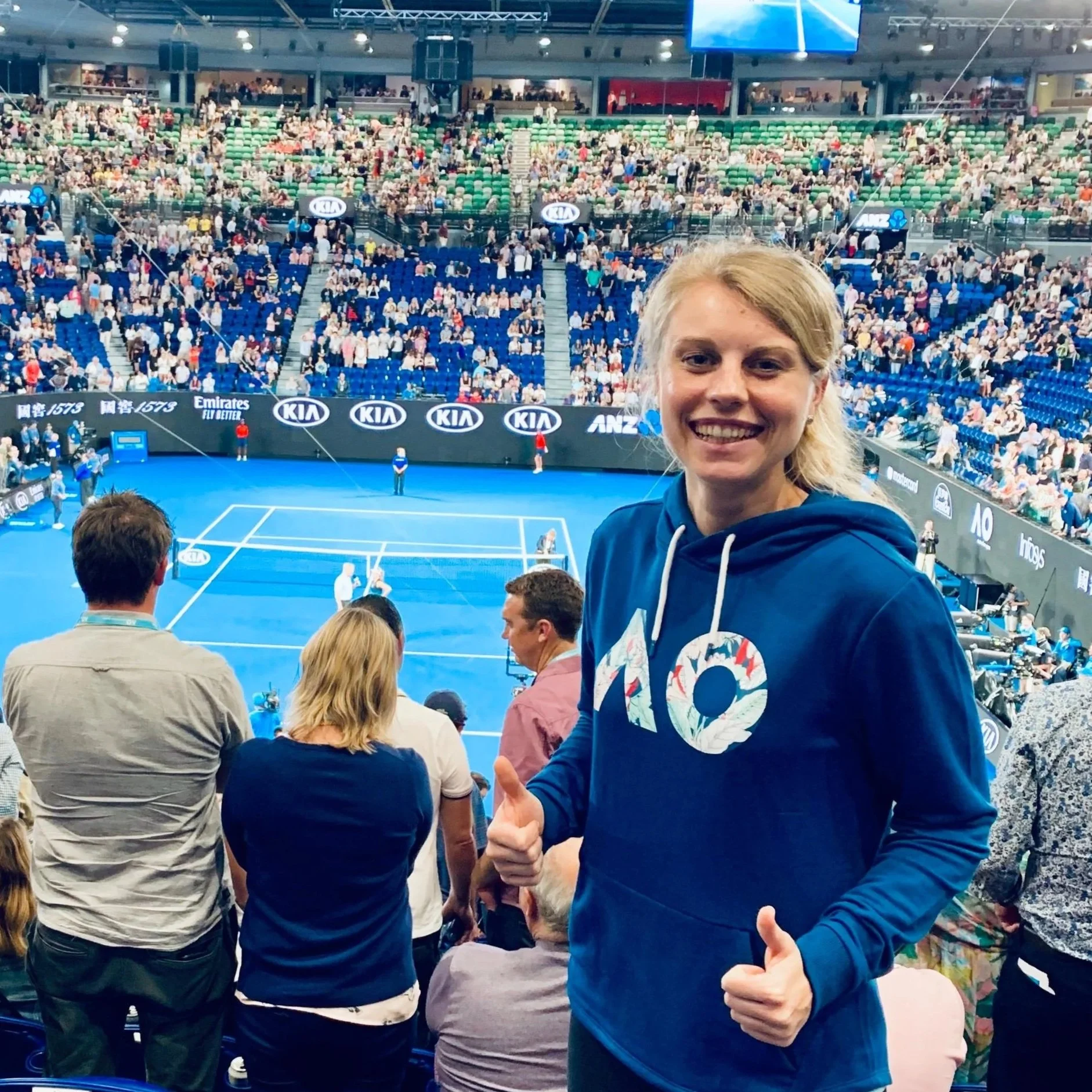 Junge Frau zeigt Daumen hoch bei einem Tennismatch in einer großen Halle, gefüllt mit Zuschauern. Sie trägt eine dunkelblaue Hoodie mit einem floral bedruckten Logo und lächelt in die Kamera.