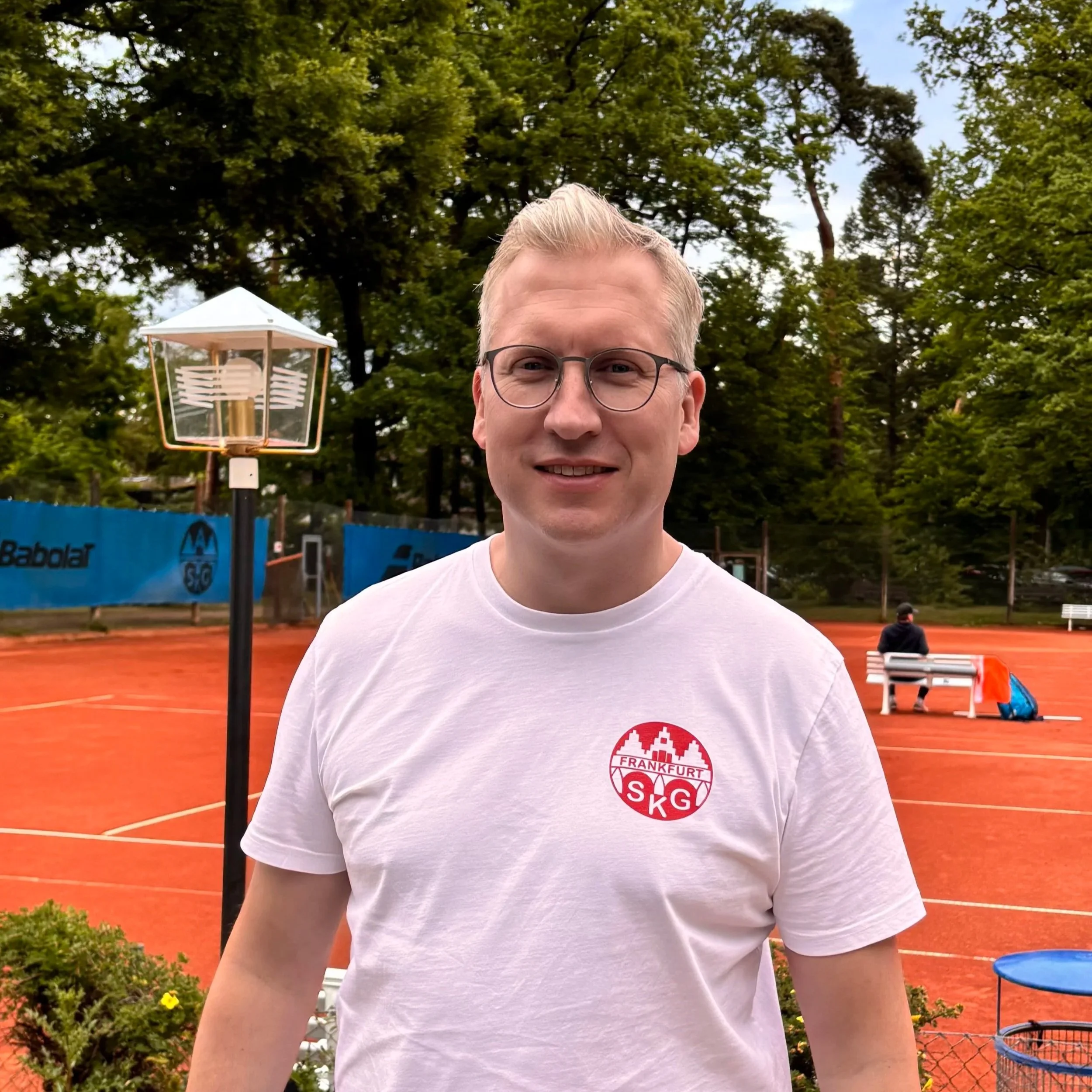 Mann steht auf Tennisplatz mit Tennisnetz, Bällen und Bänken, trägt weißes T-Shirt mit 'Frankfurt SKG' Logo, im Hintergrund Bäume und ein Erwachsener sitzend auf Bank.