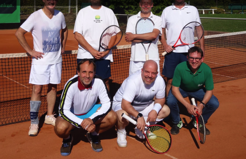 Sieben Männer auf einem Tennisplatz mit Tennisschlägern, einige stehen, einige sitzen oder knien, im Hintergrund Tennisnetz und Bäume.