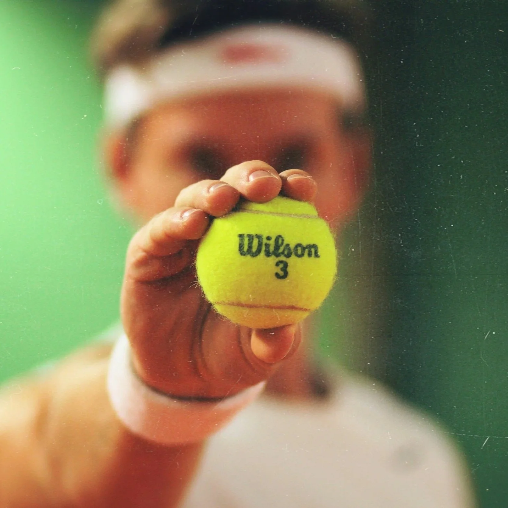 Nahaufnahme eines Tennisballs mit der Aufschrift "Wilson 3", gehalten von einer Person auf einem Tennisplatz.