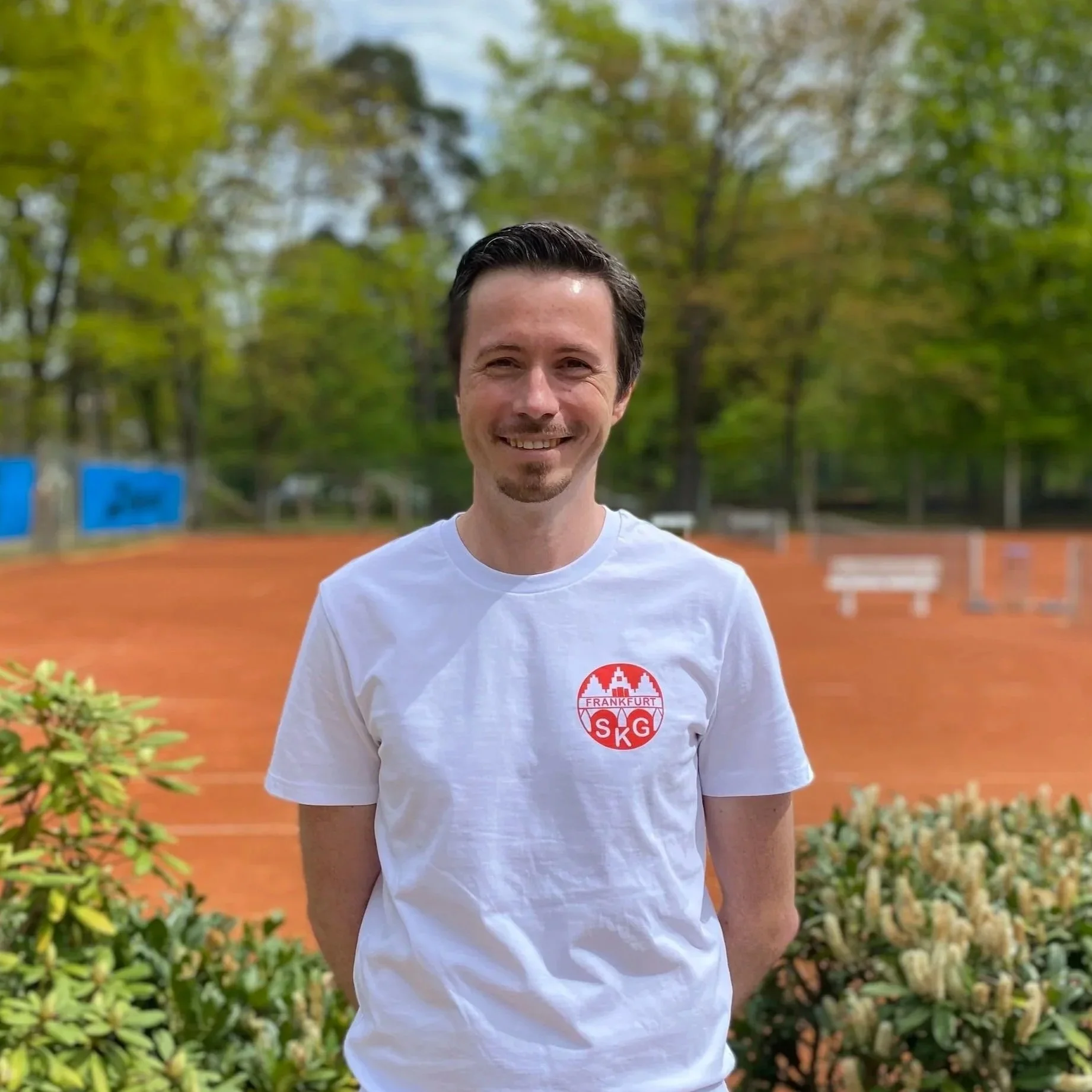 Ein lächelnder Mann steht auf einem Tennisplatz im Freien, trägt ein weißes T-Shirt mit einem roten und weißen Logo. Im Hintergrund sind Bäume und Sitzbänke sichtbar.