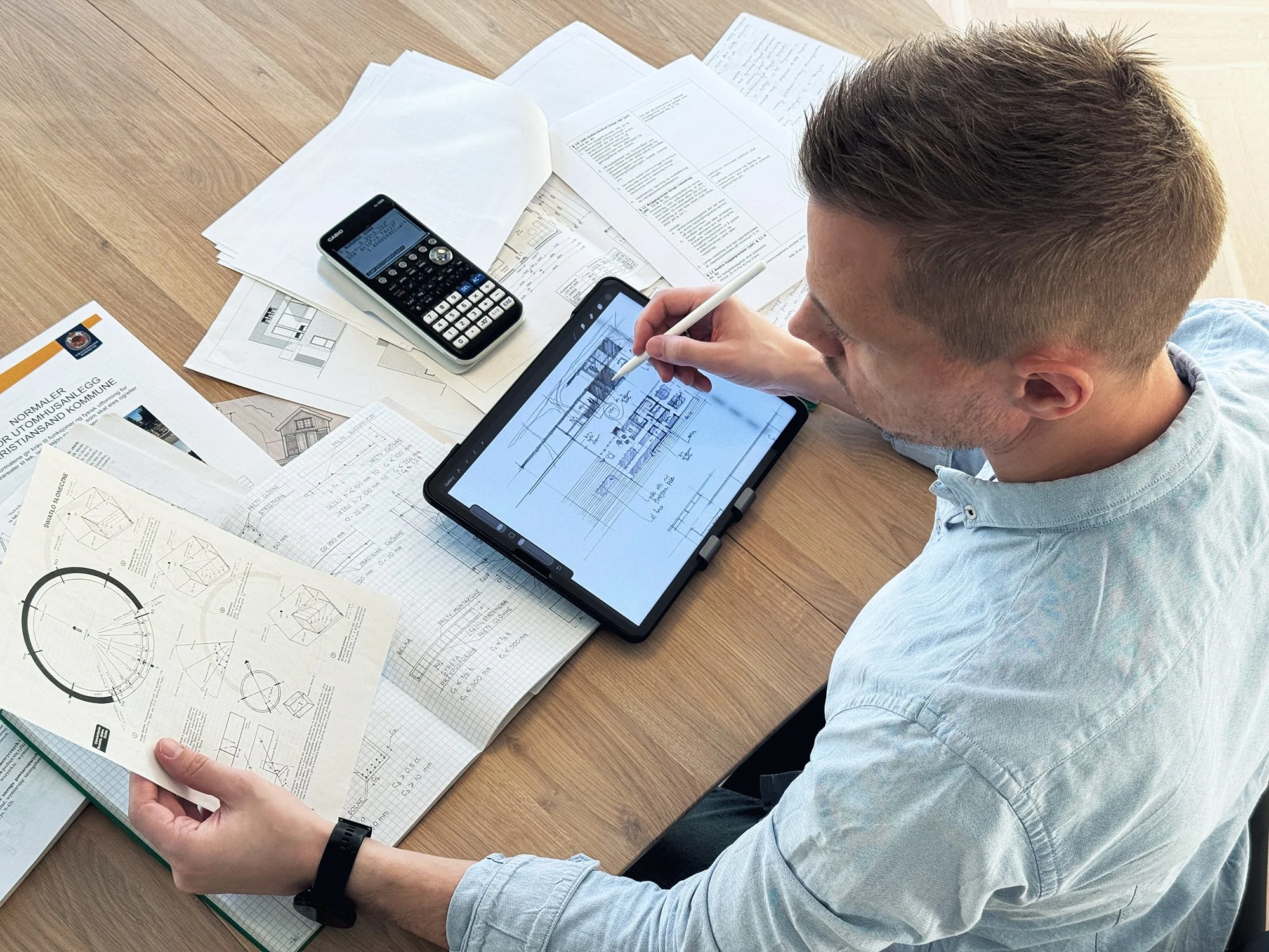 Arkitekt sitter ved bord og jobber med arkitekttegninger og skisser på iPad, omgitt av papirer og kalkulator. Bolig, tilbygg/påbygg, bruksendring, fasadeendring