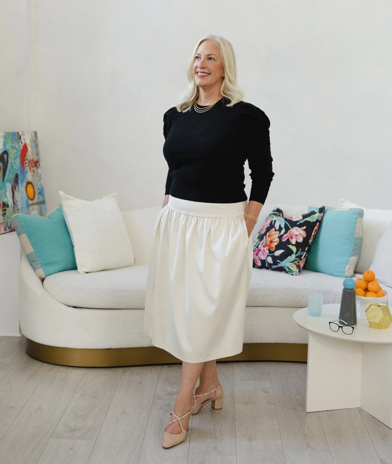A woman with blonde hair wearing a black top, cream-colored skirt, and beige heels standing in a modern living room with a white sofa, colorful pillows, a side table with items including glasses, a blue cup, and a bowl of oranges.
