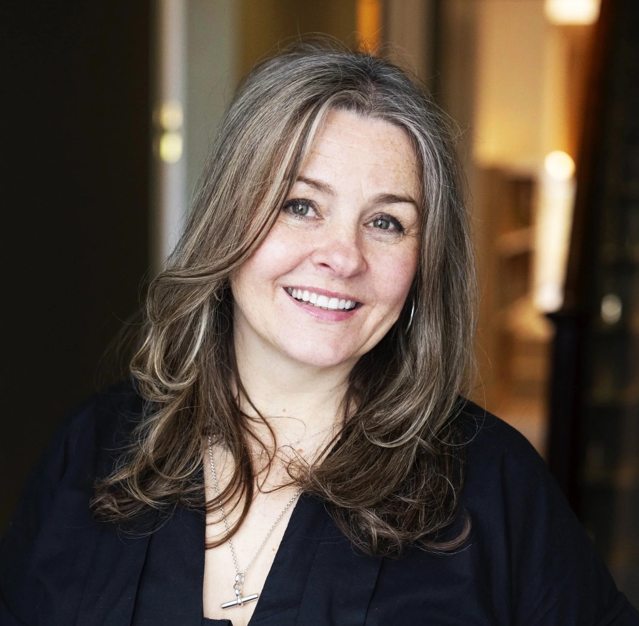 An attractive female therapist with long, wavy hair and fair skin, smiling, wearing a black top and a silver cross necklace, in an indoor setting with warm lighting.