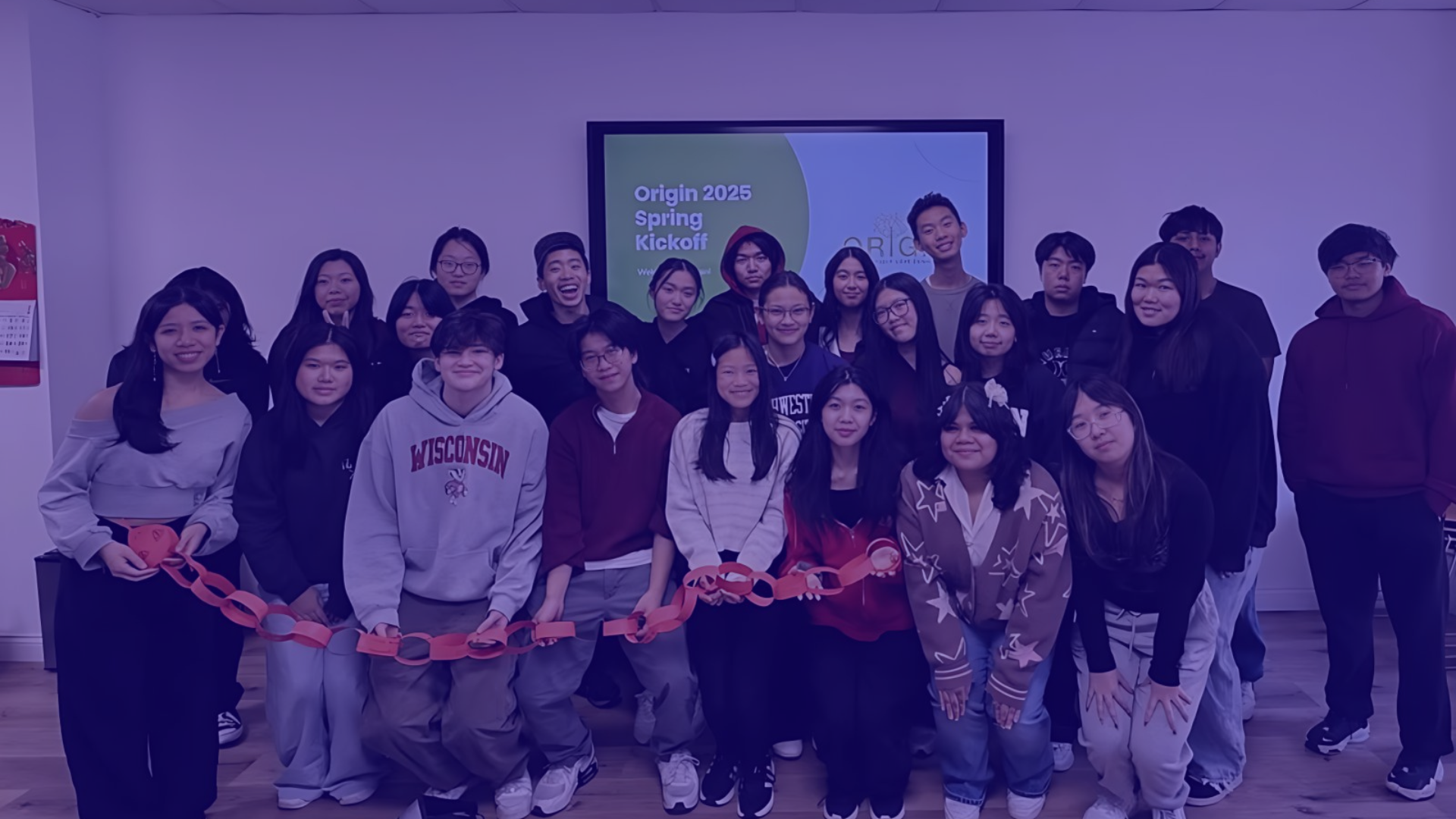 Group of young adults gathered indoors for a group photo, some holding a paper chain, with a screen in the background displaying 'Origin 2025 Spring Kickoff.'