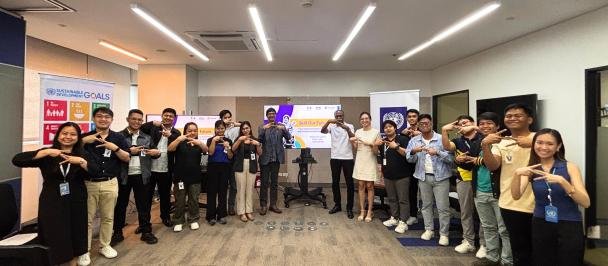 Group of diverse people standing in a conference room, holding hands in a chain, with presentation screens and banners in the background.
