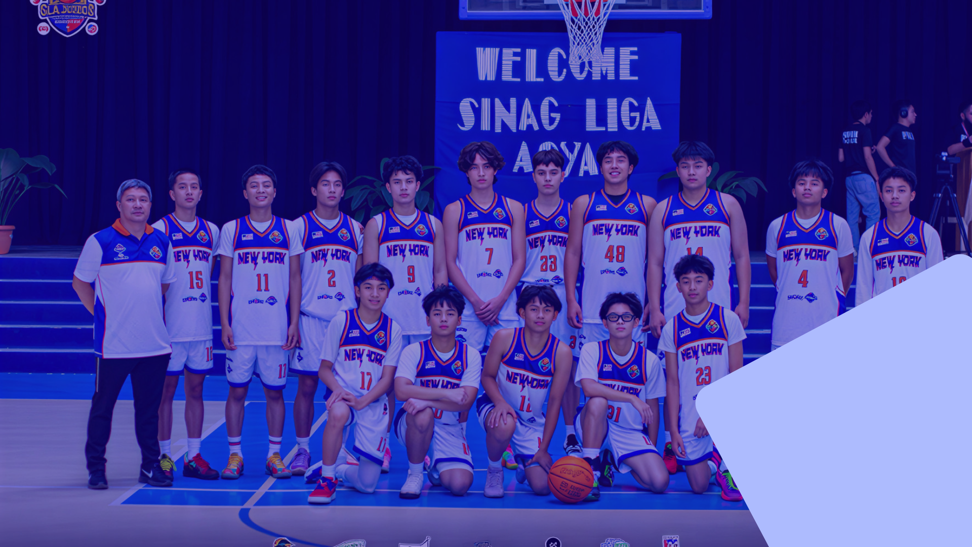 Group of young basketball players and coach posing for a team photo on a basketball court. They are wearing white and blue jerseys with 'New York' and various numbers. A sign behind them reads 'Welcome Sinag Liga'.