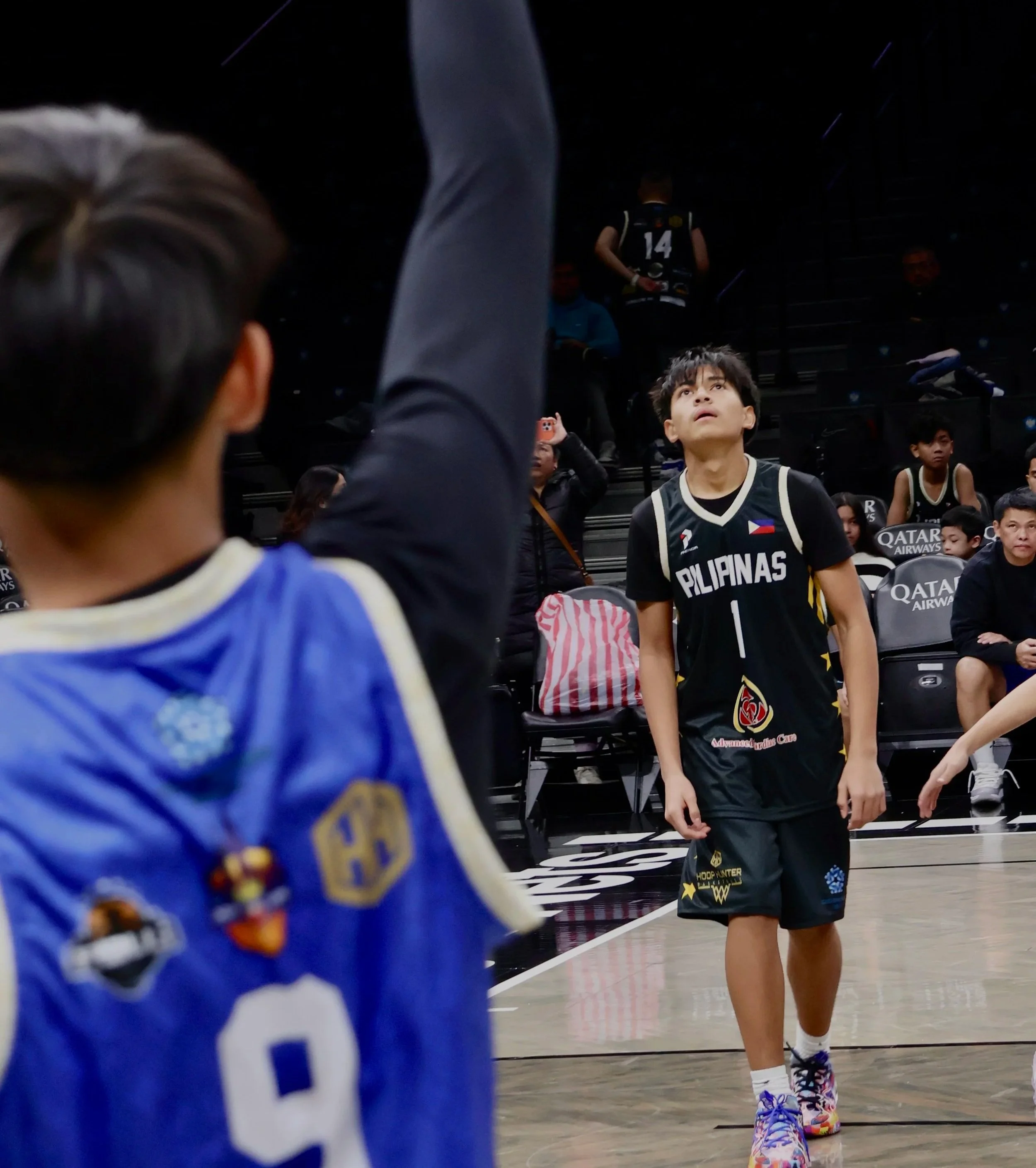 Young boy in basketball uniform with 'Pilipinas' written on it and the number 1, standing on a basketball court during a game, looking upwards with focus. Other players and spectators are visible in the background.
