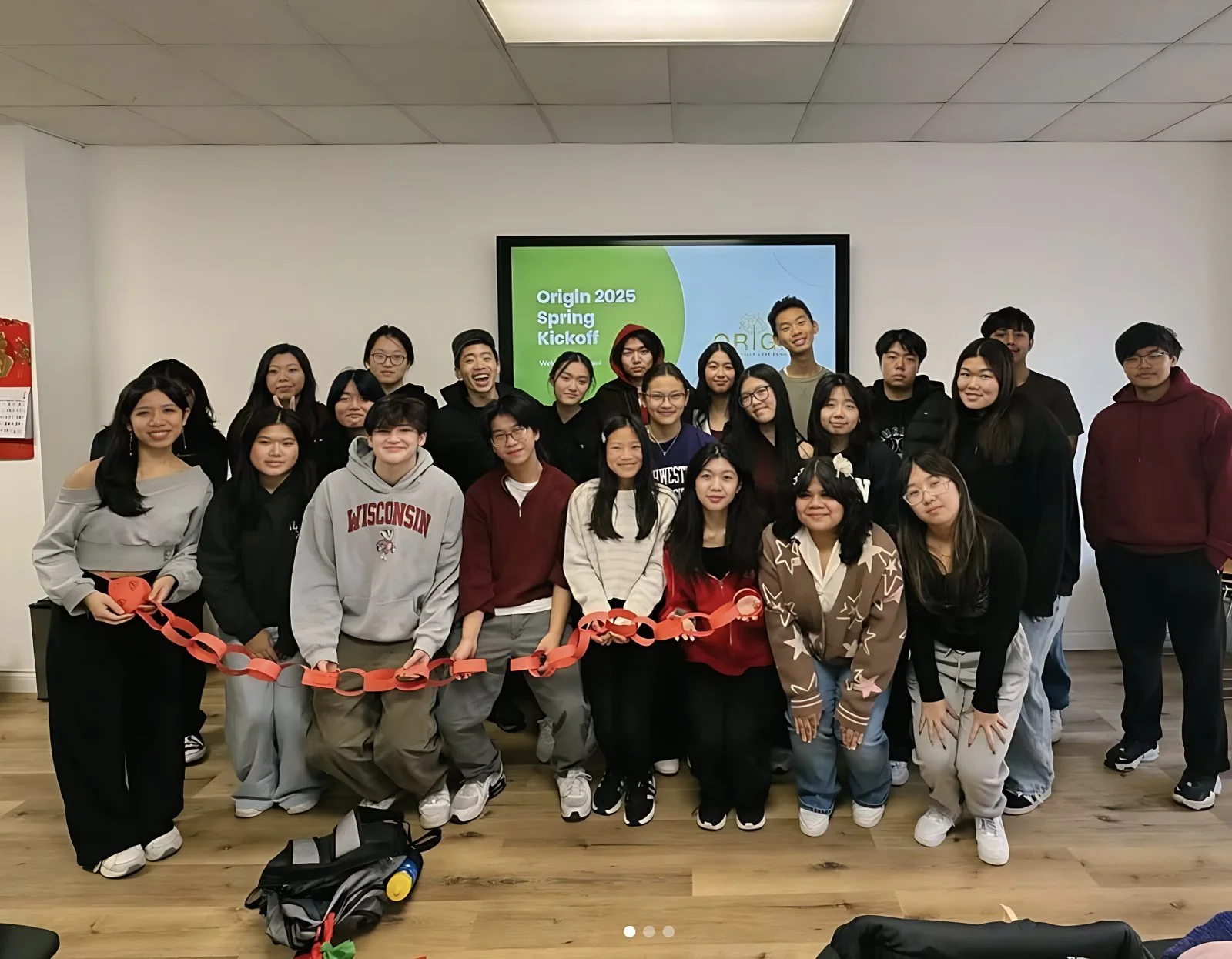 Group photo of students gathered in a classroom for the Origin 2025 Spring Kickoff event, with a screen displaying the event title in the background, and the students holding a paper chain.