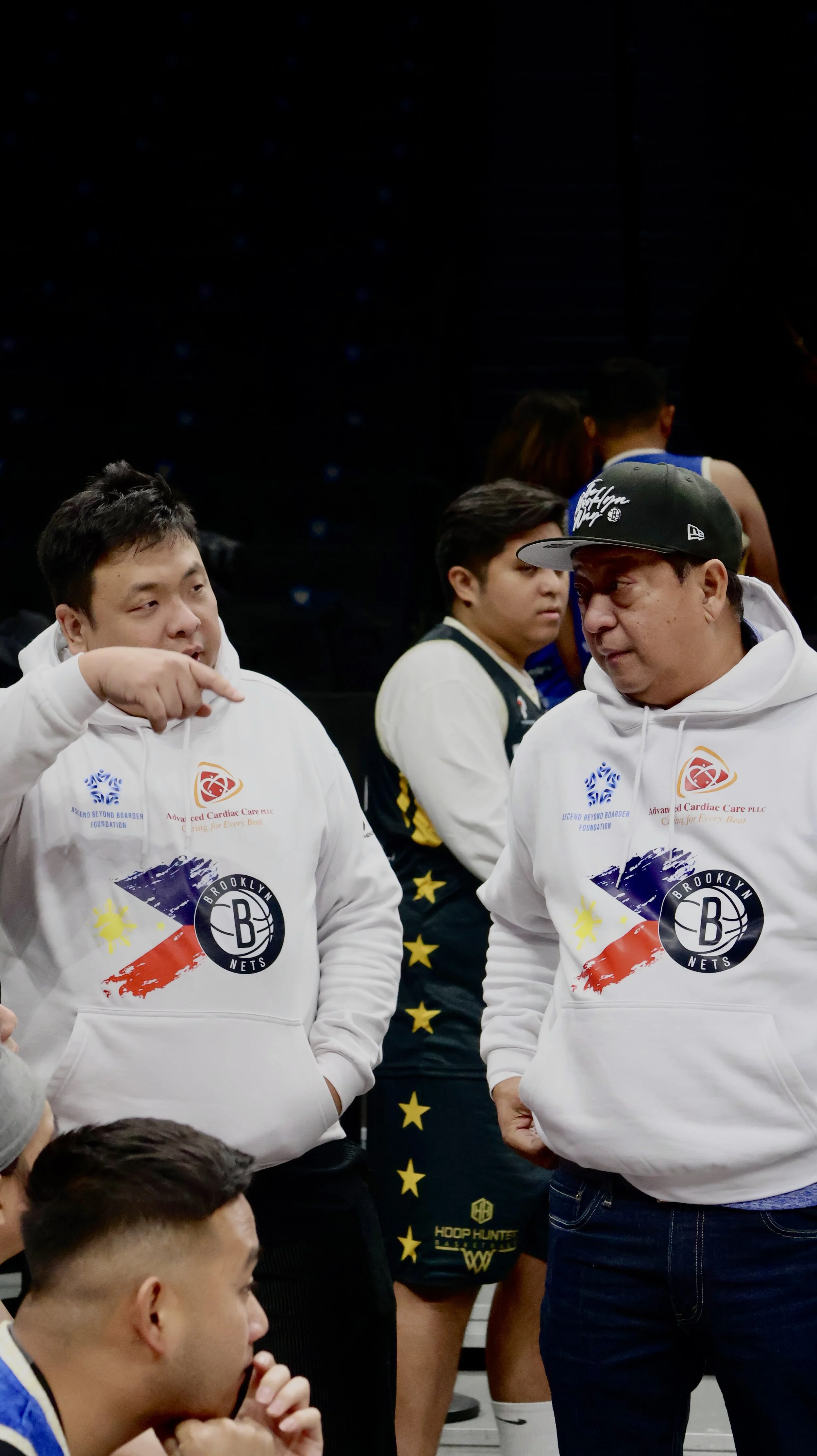 Two men in Brooklyn Nets hoodies talking to each other among basketball players and other people in the background.