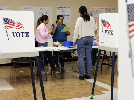 A woman at a table voting with election officials and voting booths decorated with American flags in the background.
