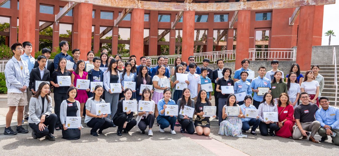 A large group of young adults and teenagers posing outdoors, holding certificates, in front of a red brick building.