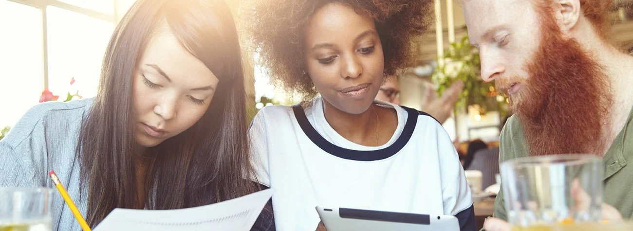 Three diverse young adults sitting at a table in a cafe, looking at a tablet and printed papers, engaged in discussion.