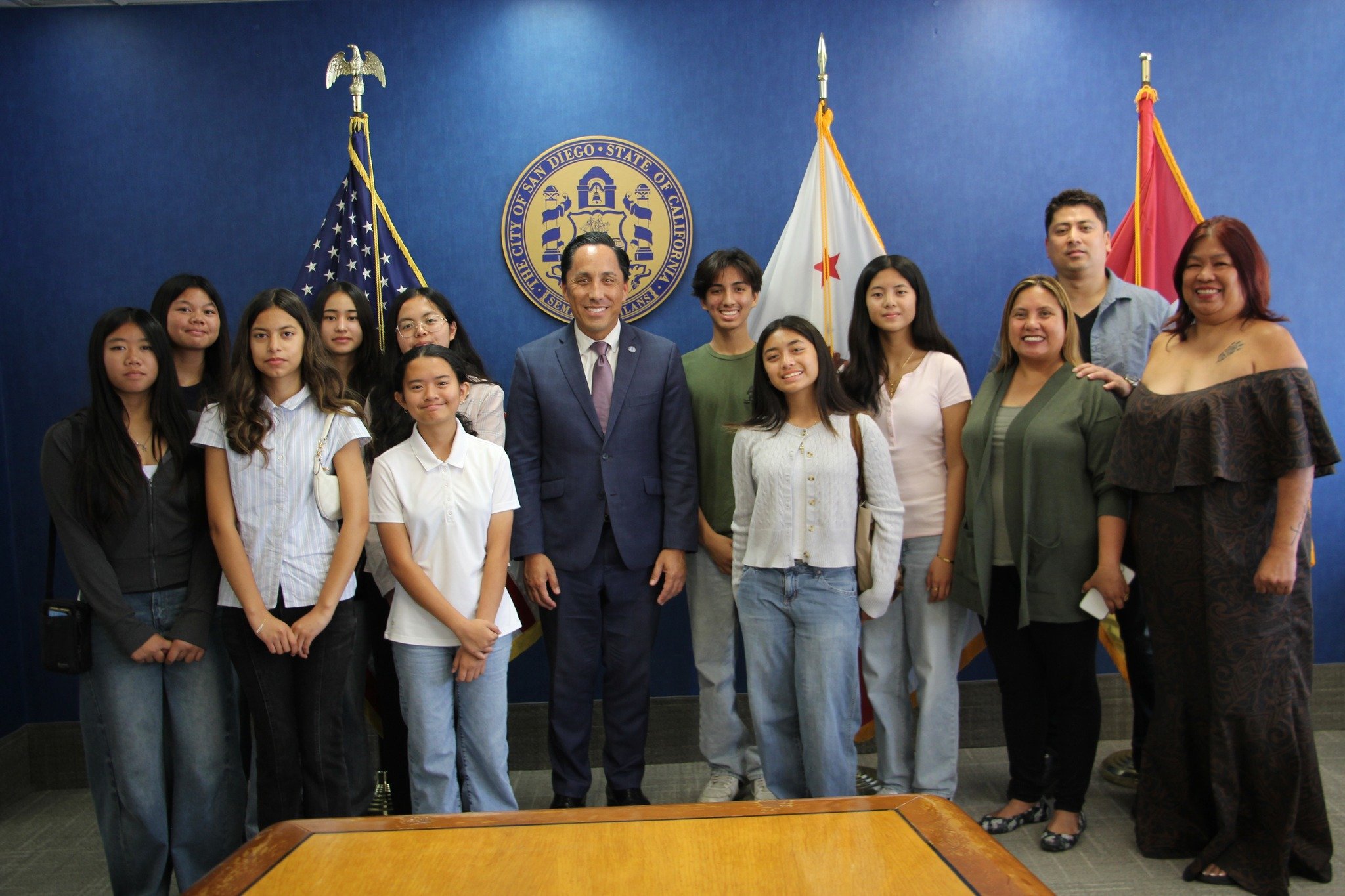 Group photo of people standing in front of a blue wall with the seal of the City of San Diego, California, flags, and smiling for the camera.