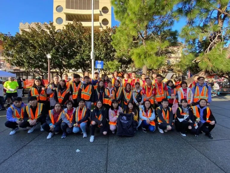 Group of young people wearing orange safety vests posing outdoors in front of trees and a building.