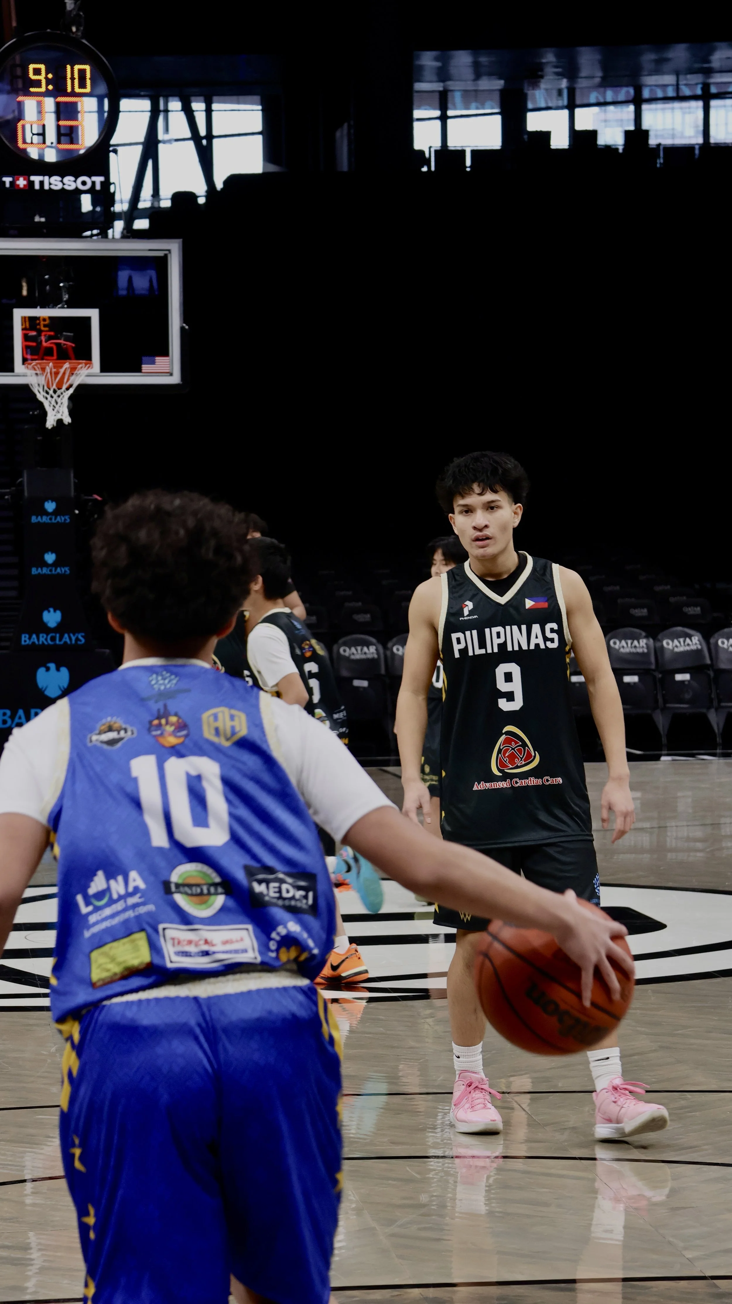 A young male basketball player wearing a black jersey with 'PILIPINAS' and the number 9 dribbling a basketball on an indoor court, with another youth in a blue jersey with the number 10 facing him.