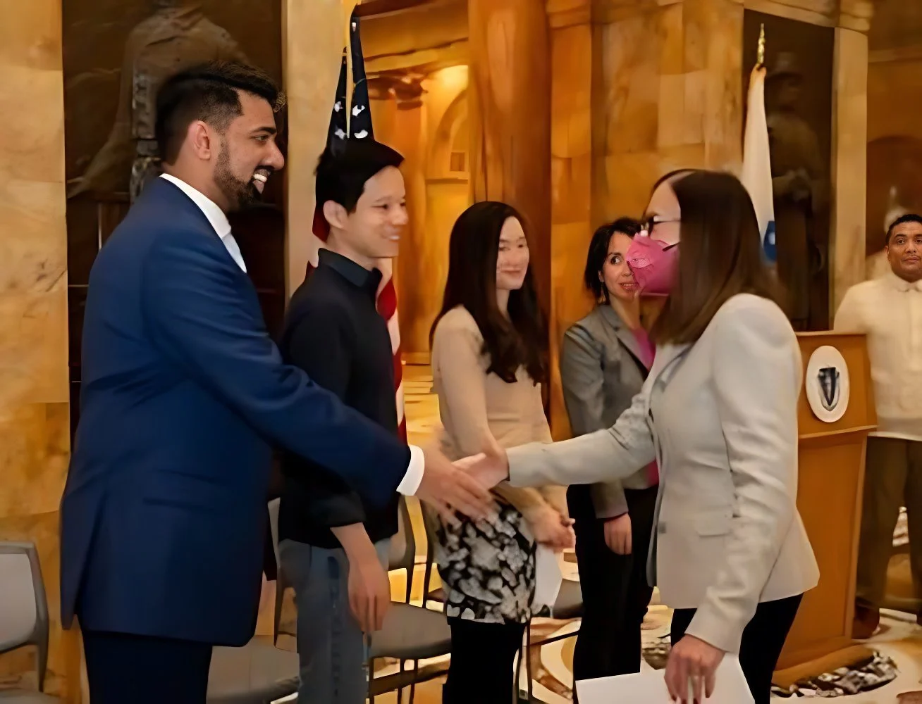 Group of people at a formal event, shaking hands, with one woman wearing a pink face mask.