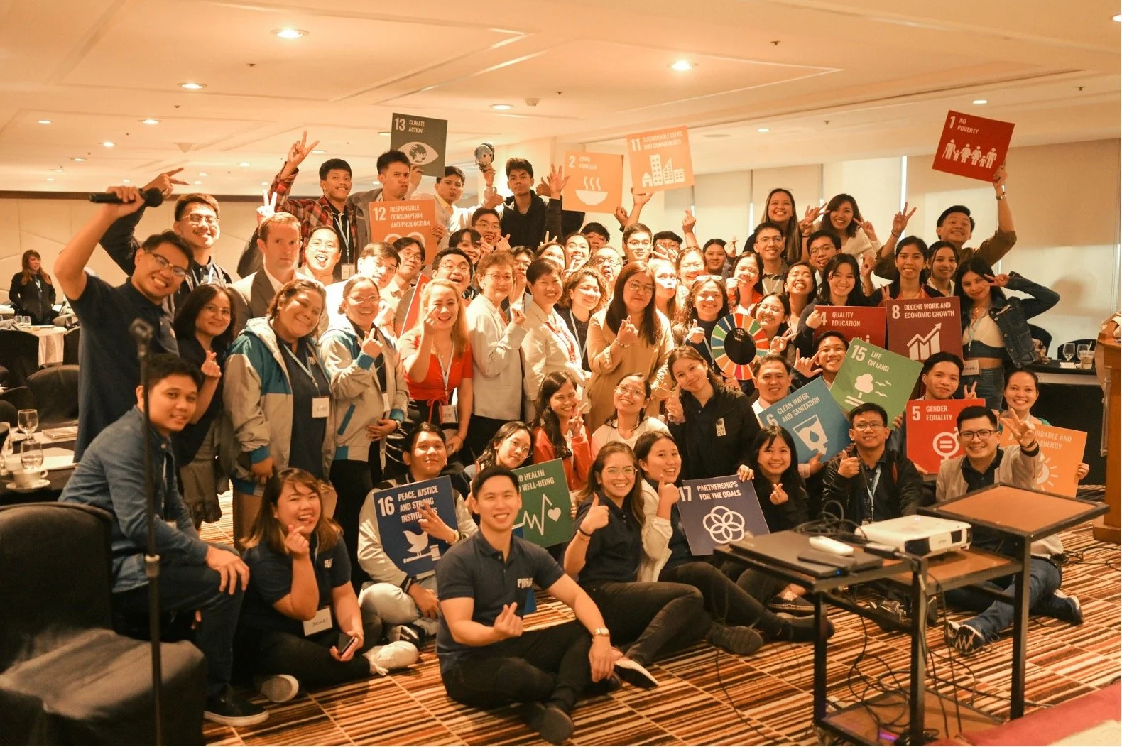 A large group of diverse people gathered in a conference room, smiling and holding signs representing various United Nations Sustainable Development Goals.