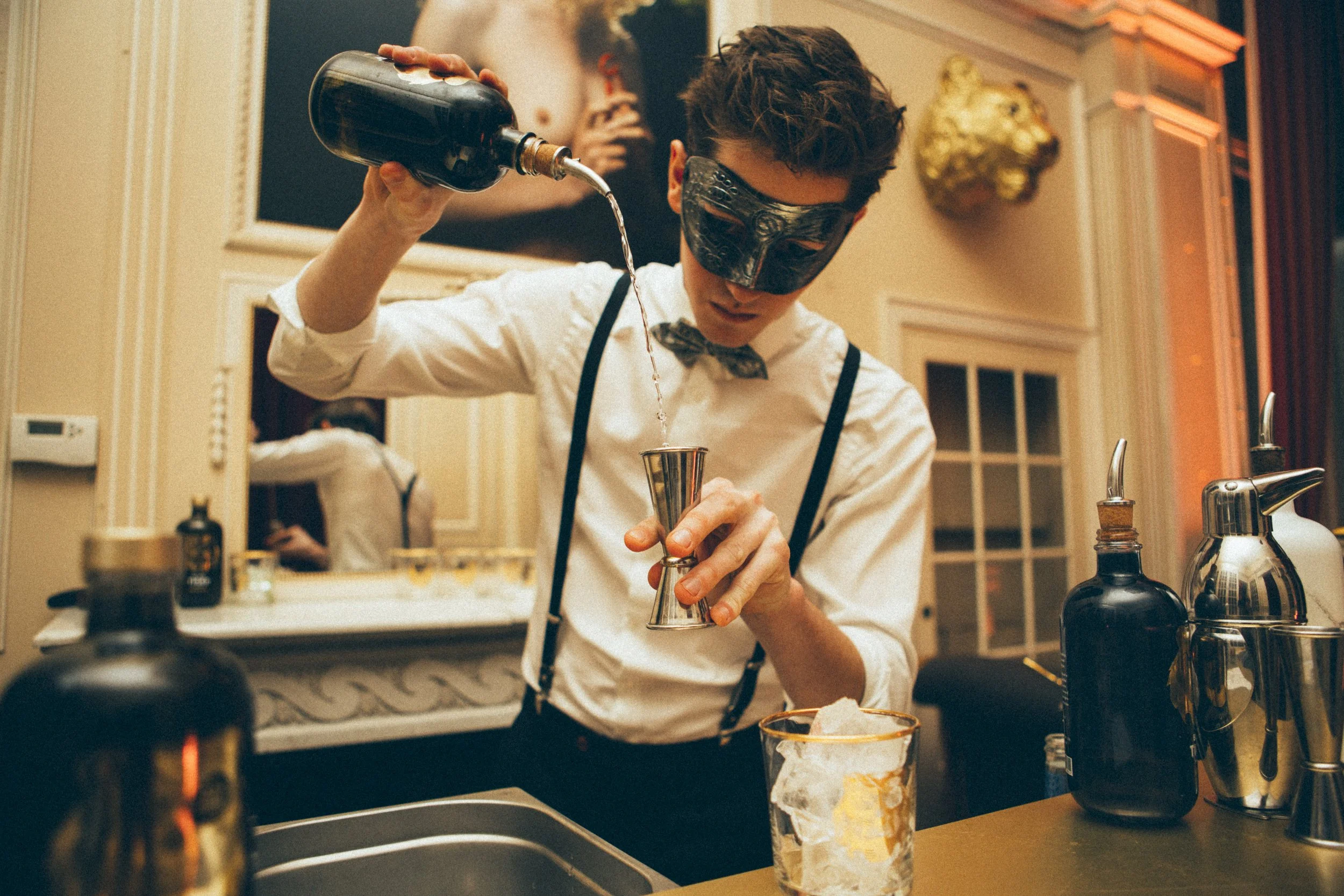 Person in mask pouring liquid into a jigger at a bar, with various bar tools and bottles on the counter.
