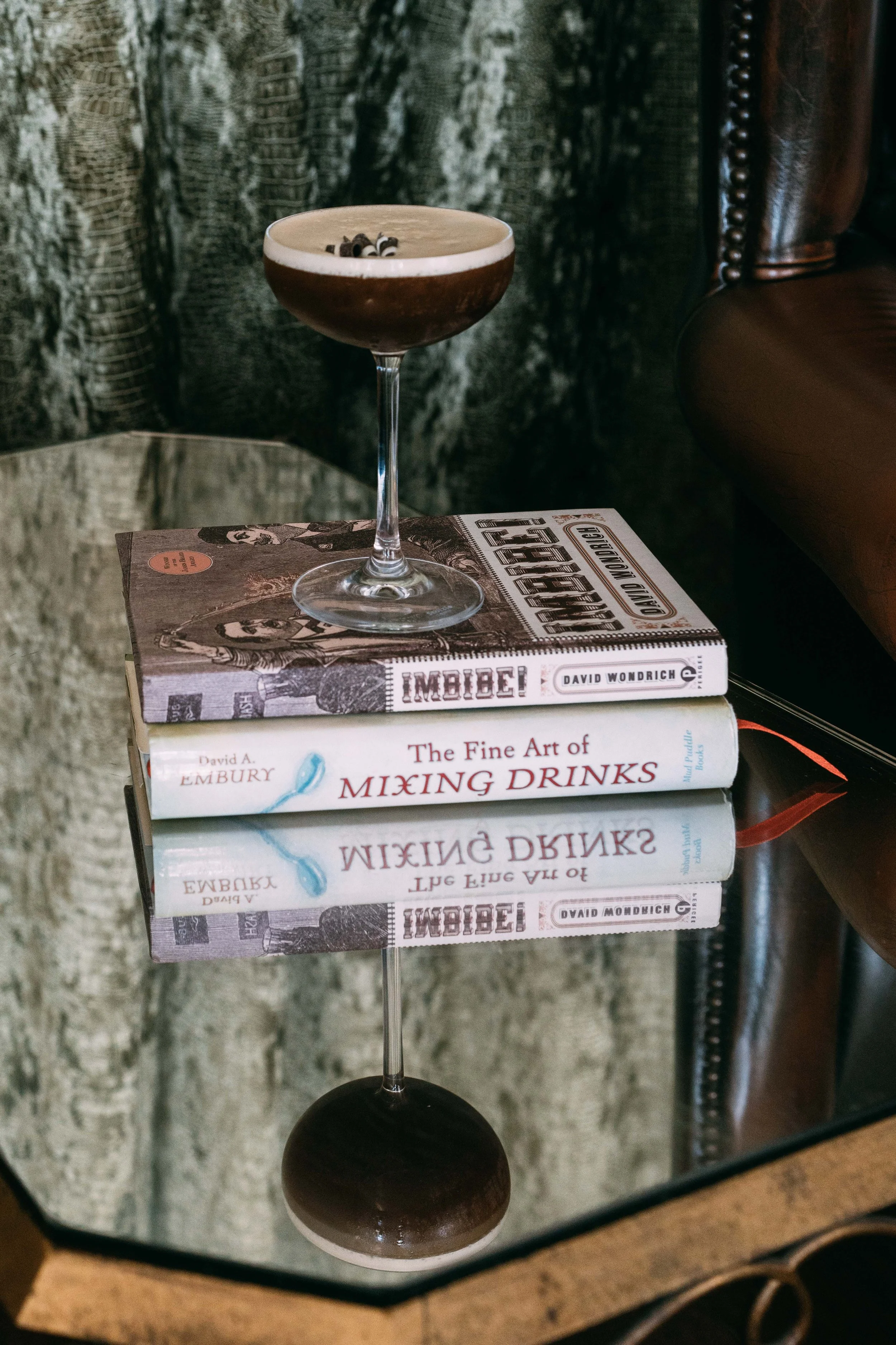 A black and white cocktail in a coupe glass with chocolate shavings on top, placed on top of three stacked books on a glass table, with a mirror reflecting the books and drink.