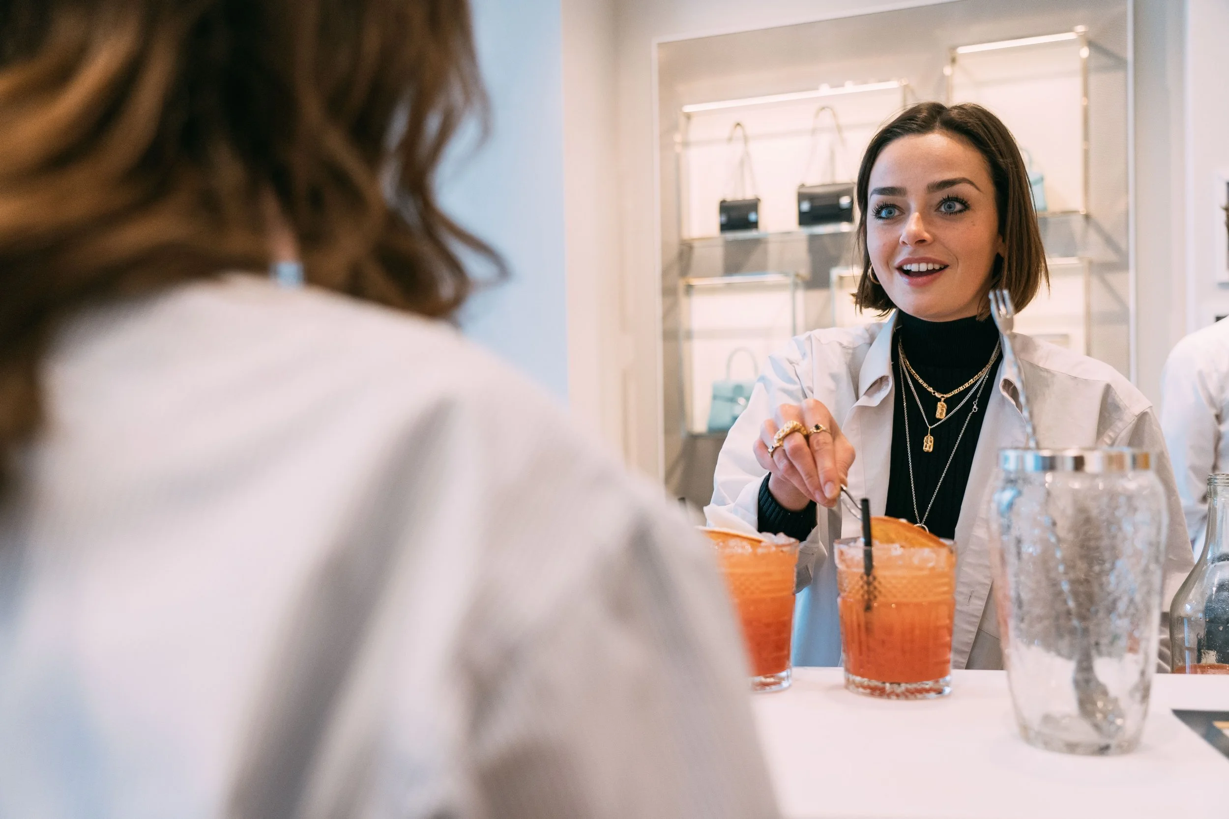 A woman with short dark hair and layered necklaces, wearing a black turtleneck and white shirt, smiles and stirs drinks at a table while talking to another woman with long brown hair, in a retail store.