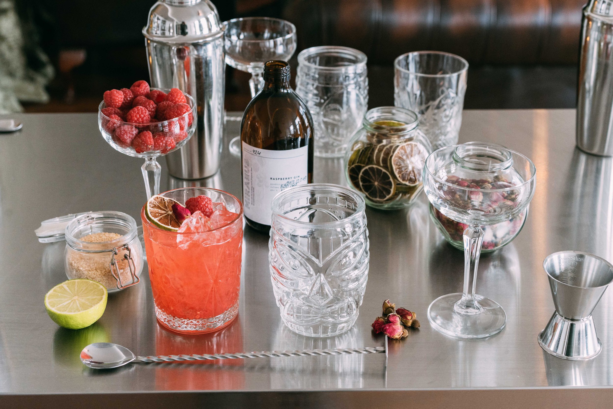 A stainless steel bartop with various cocktail-making ingredients, including fresh raspberries, dried lemon slices, a halved lime, a jar of sugar, a bottle of raspberry gin, and glassware such as a tiki mug, coupe glasses, and a hurricane glass. There are cocktail tools like a muddler and a cocktail jigger.