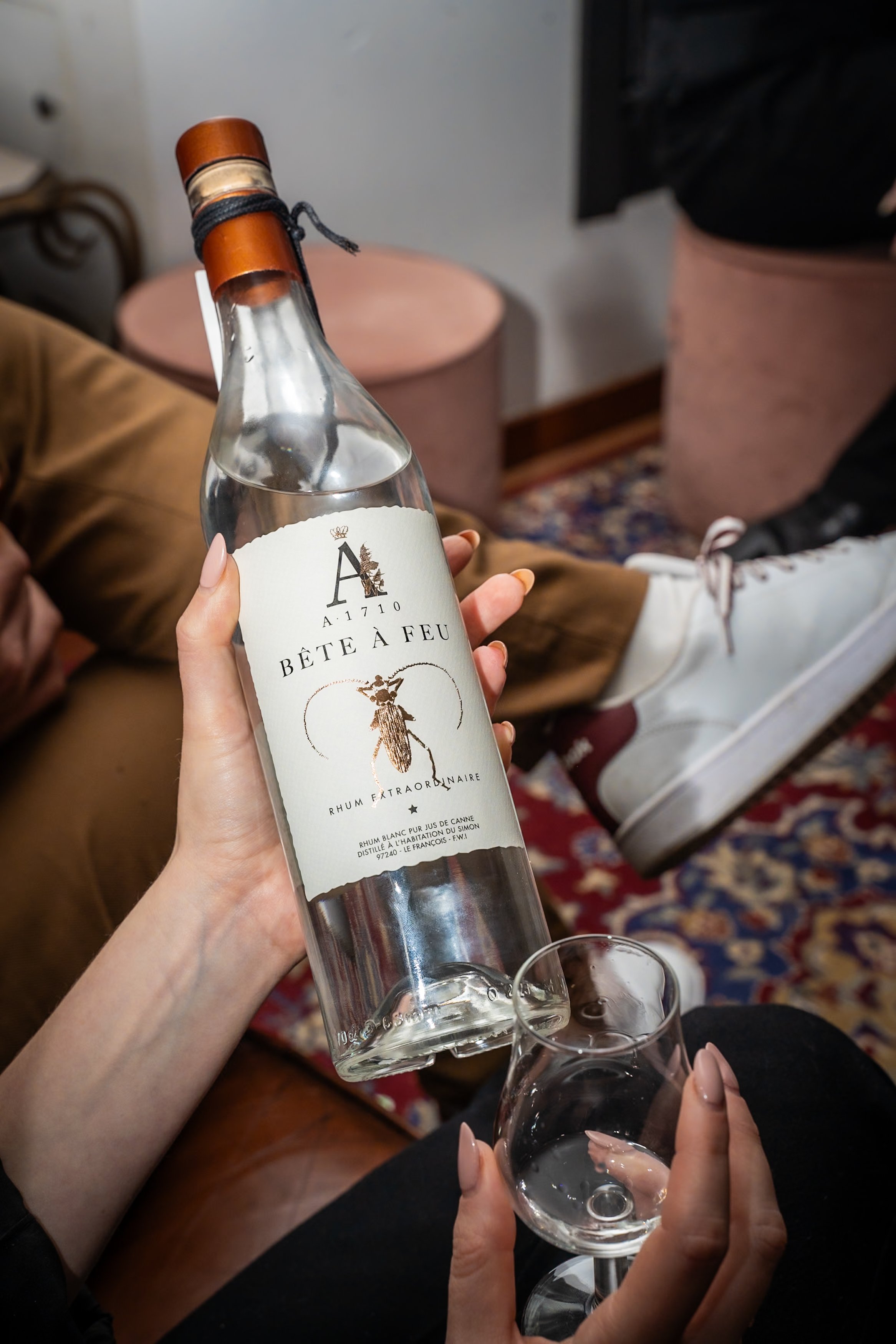 A hand holding a bottle of Bête à Feu rum and a glass of clear liquid, with a person wearing white sneakers in the background.