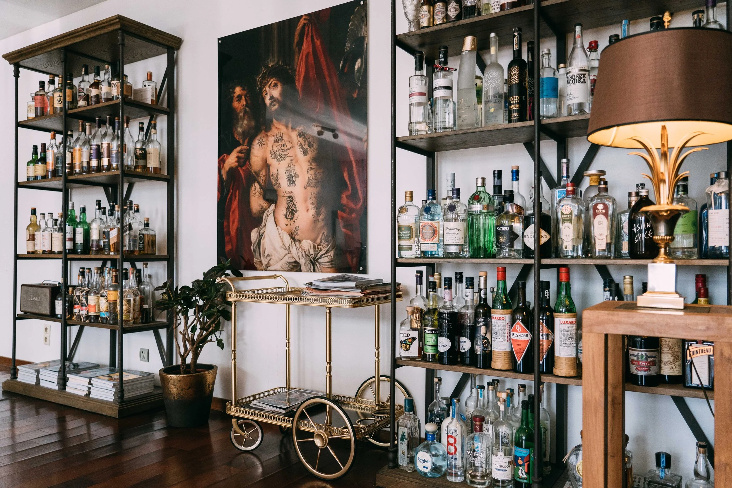 A bar corner with alcohol bottles on black metal shelves, a large painting of a tattooed man with a red cape, a potted plant, a brass bar cart, and a lamp with a brown shade.