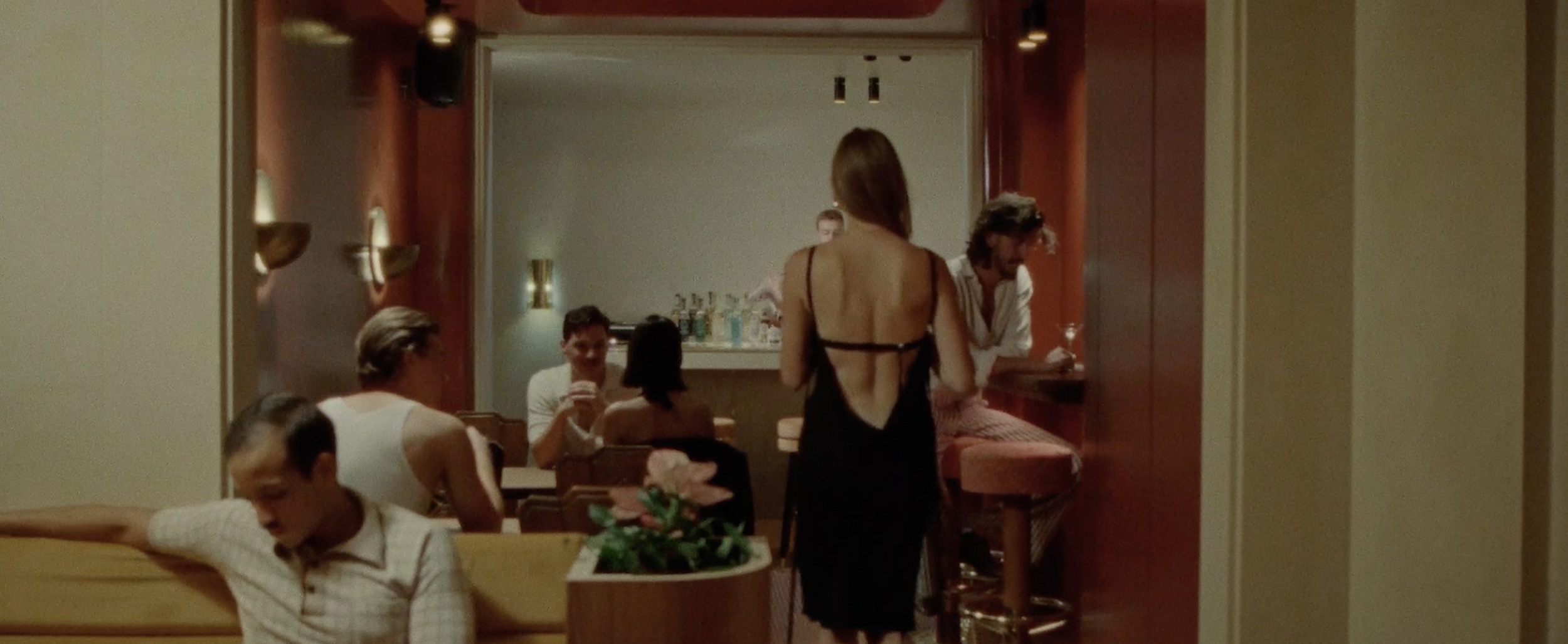 A group of people socializing at a bar or lounge, some seated and some standing, with drinks in hand, in a warmly lit interior.