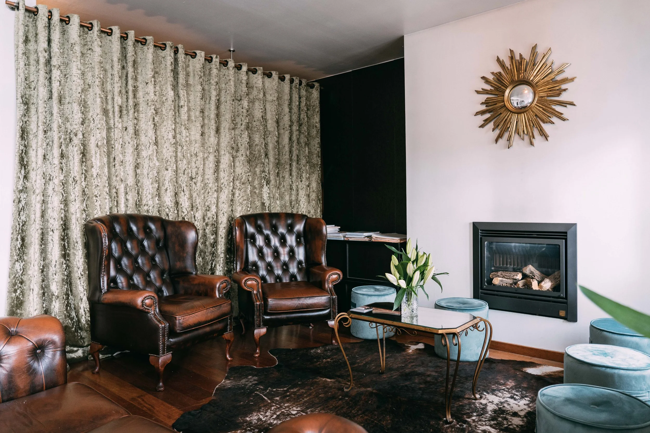 Cozy living room with leather armchairs, a marble coffee table, and a fireplace on a white wall. Gold sunburst mirror decorates the wall, and teal ottomans surround the area. Beige curtains hang beside a dark wall.