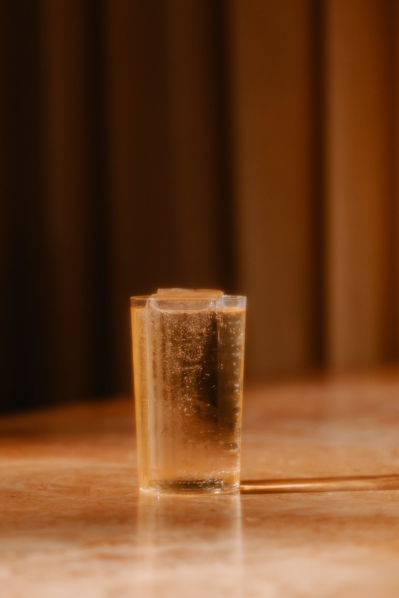 A glass with a cocktail, with ice cubes on a wooden surface with a dark, blurred background.