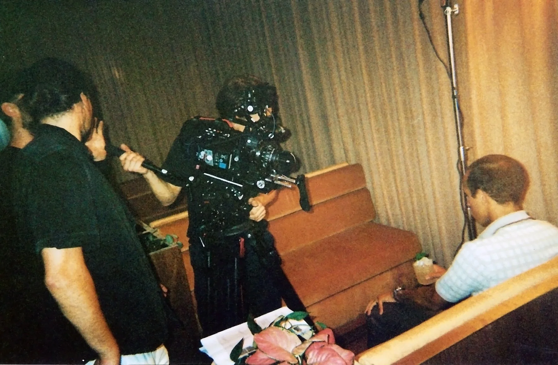 A film crew is filming a man sitting on a wooden bench in a room with curtain-covered walls. The crew includes a person holding a camera on a rig and another person holding a microphone.
