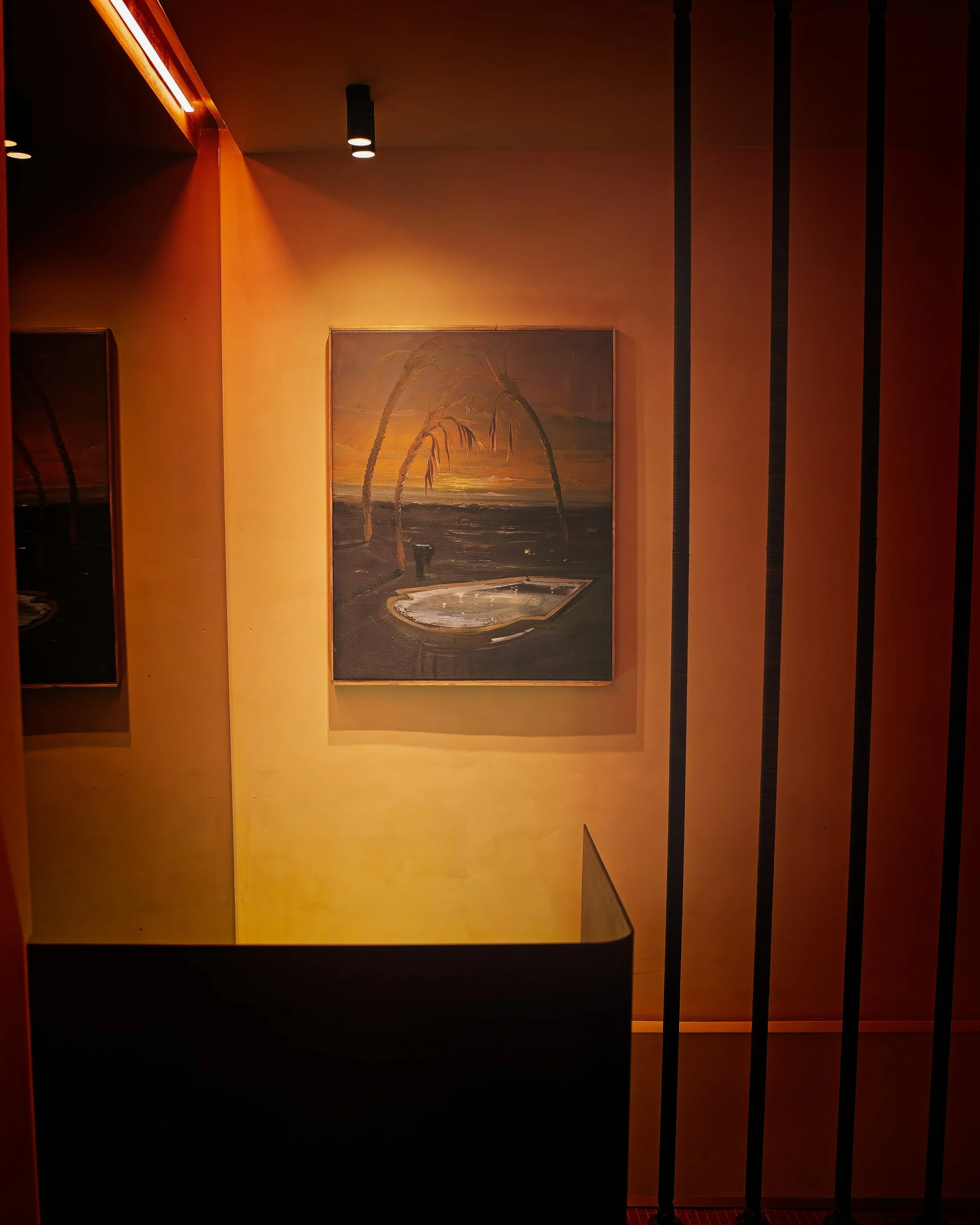 A painting of two palm trees at sunset over a body of water, with a small pond and a figure near it, displayed on a warm-colored wall in an art gallery.